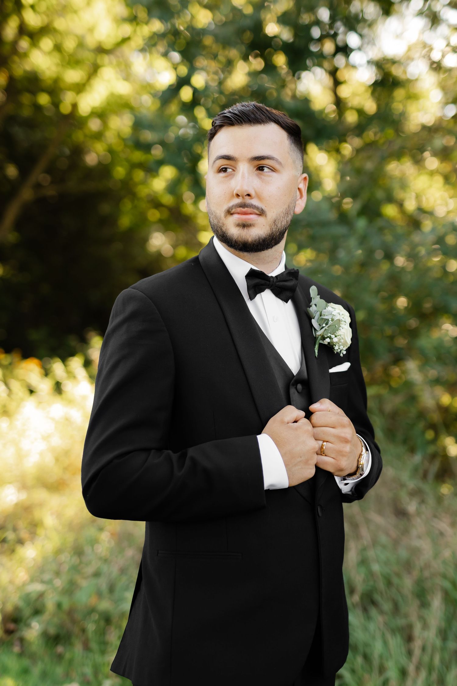 Groom in classic black tuxedo with boutonniere poses against natural outdoor backdrop.