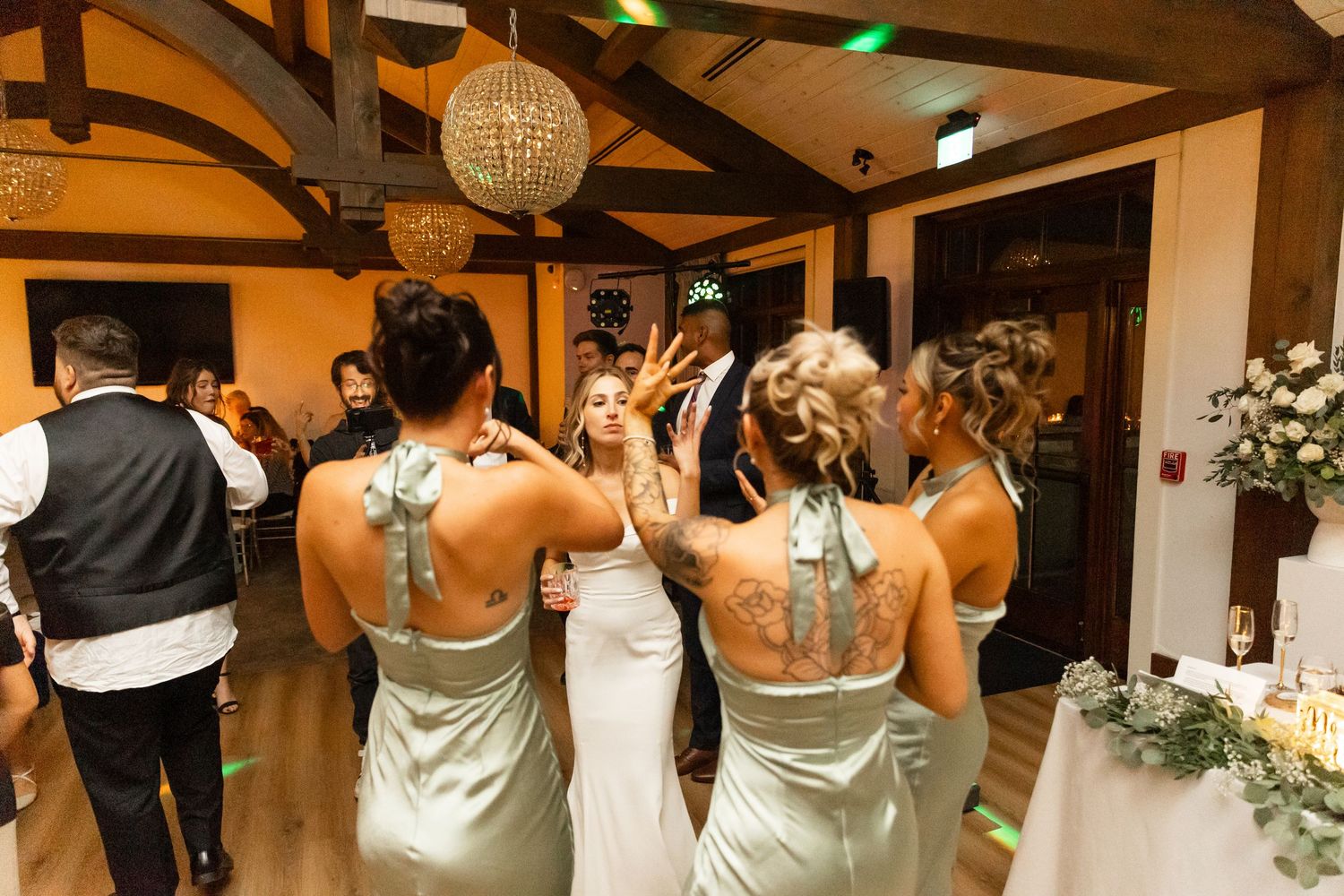 Bridesmaids in matching dresses dancing at a wedding reception under decorative lighting.