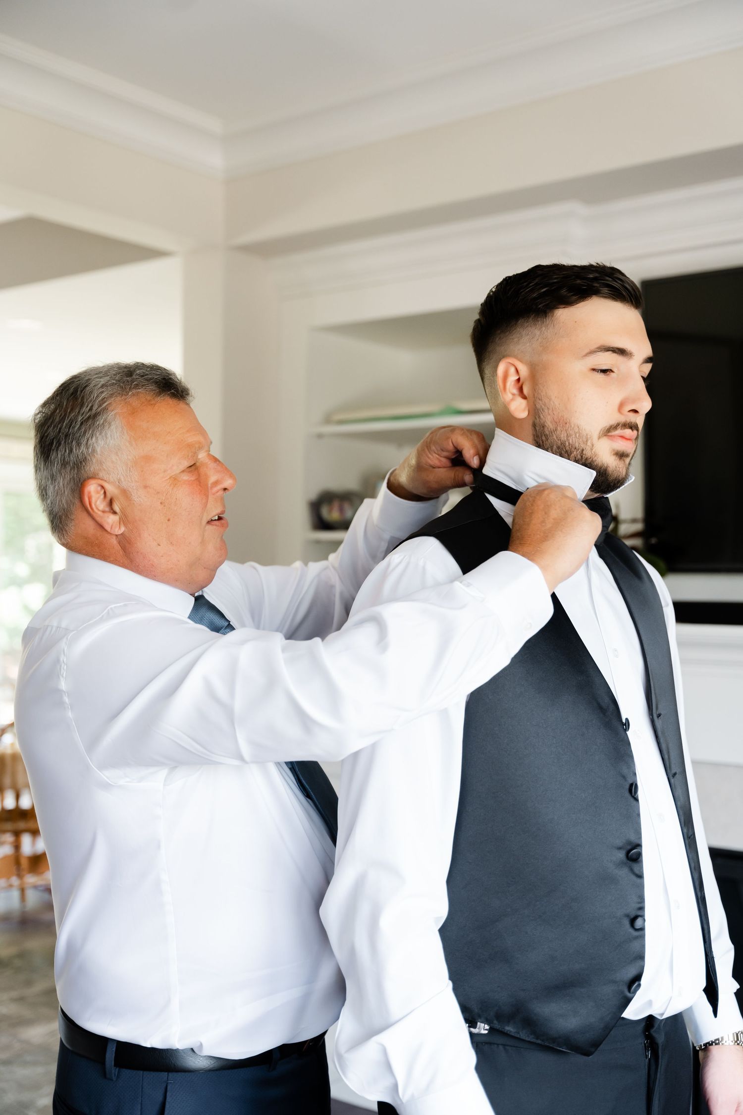 Men in white dress shirts and vests help each other prepare for a formal event.