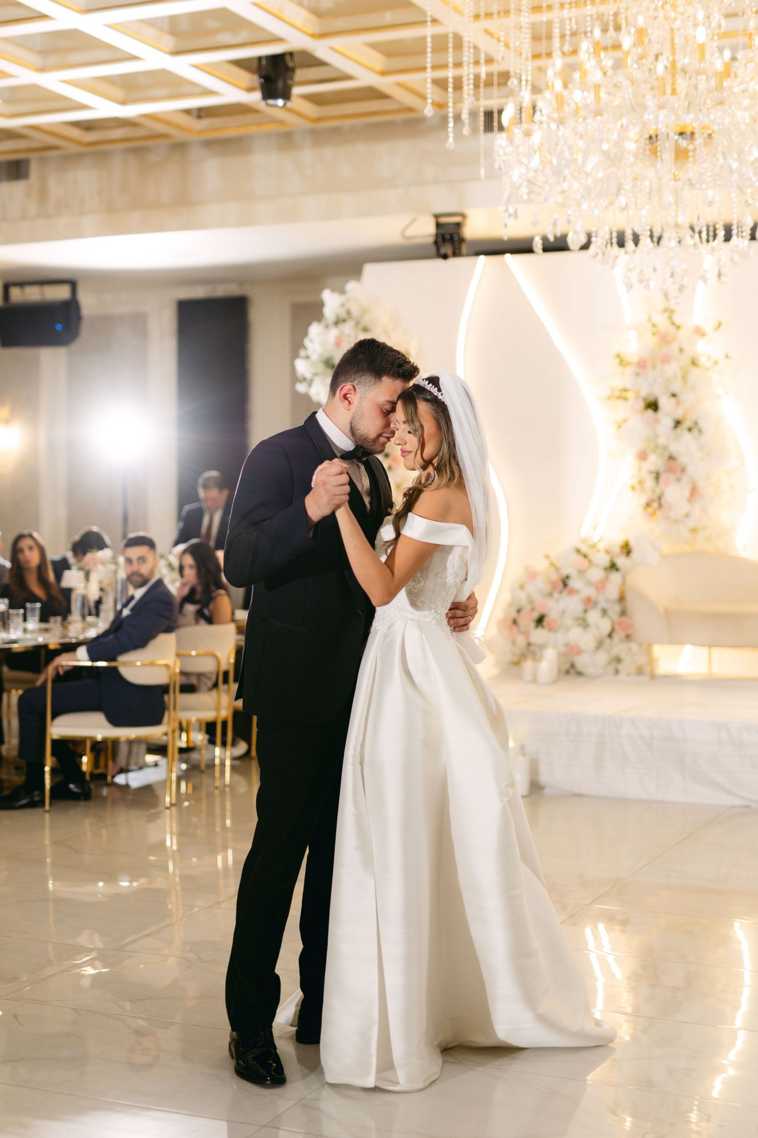 Newlyweds share their first dance in an elegant ballroom with crystal chandeliers and floral decorations.