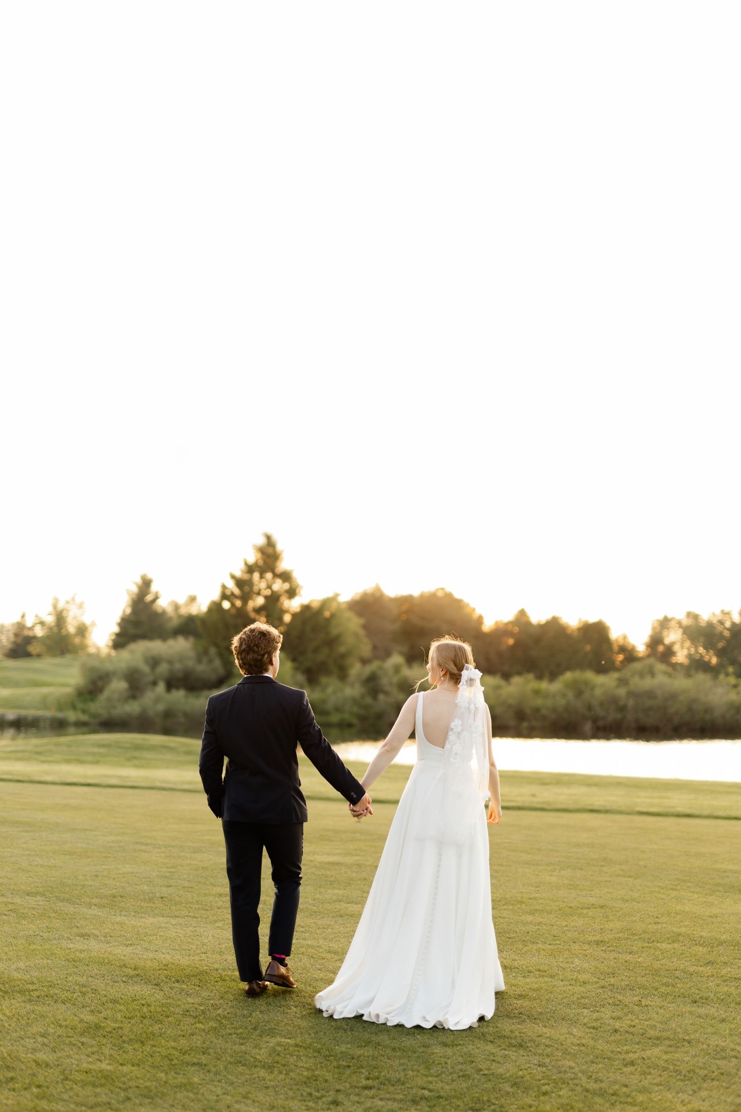 Wedding couple walking across a sunlit grassy field at golden hour.
