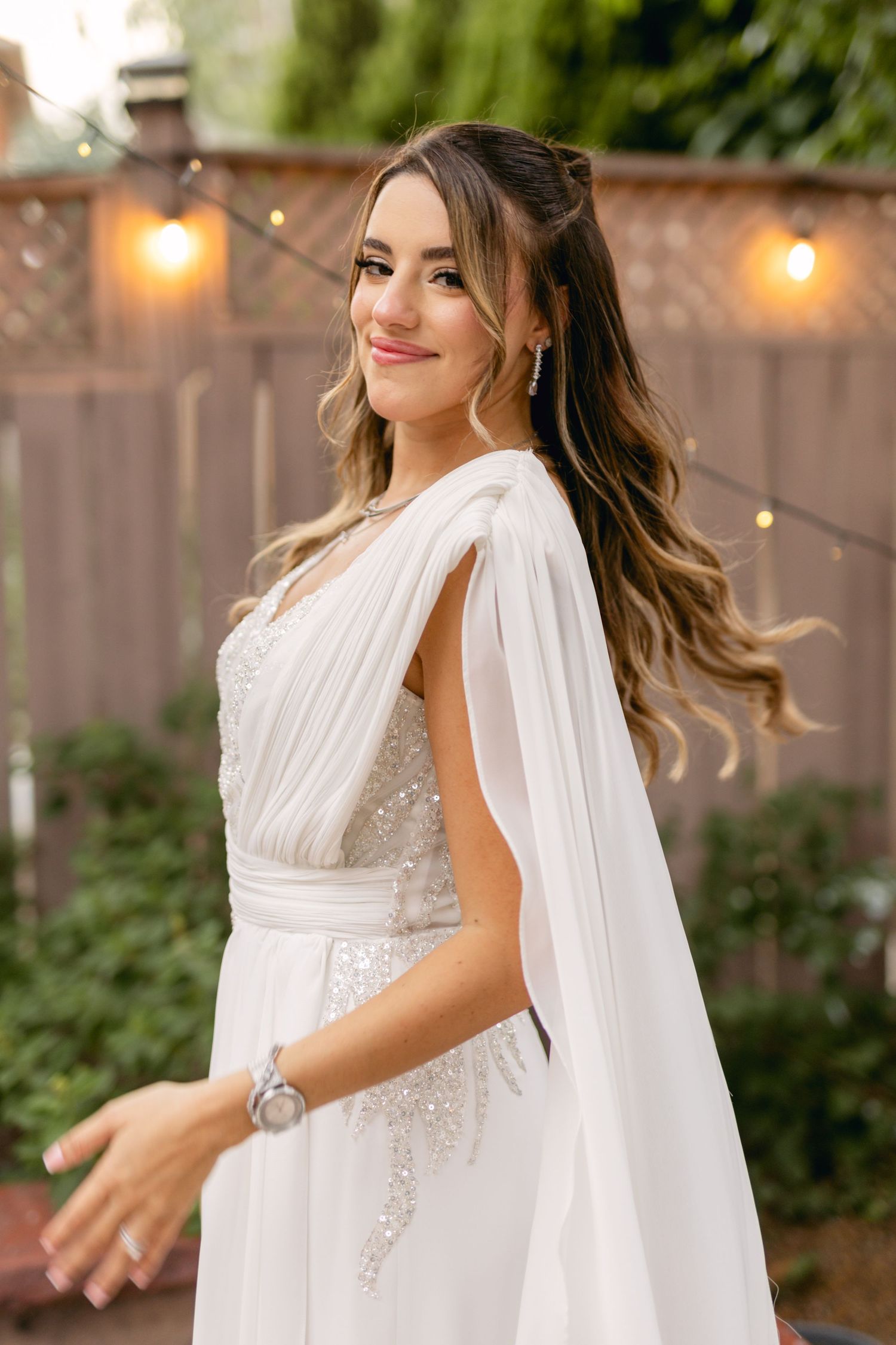 Side profile of bride in flowing white dress with cape against garden backdrop.