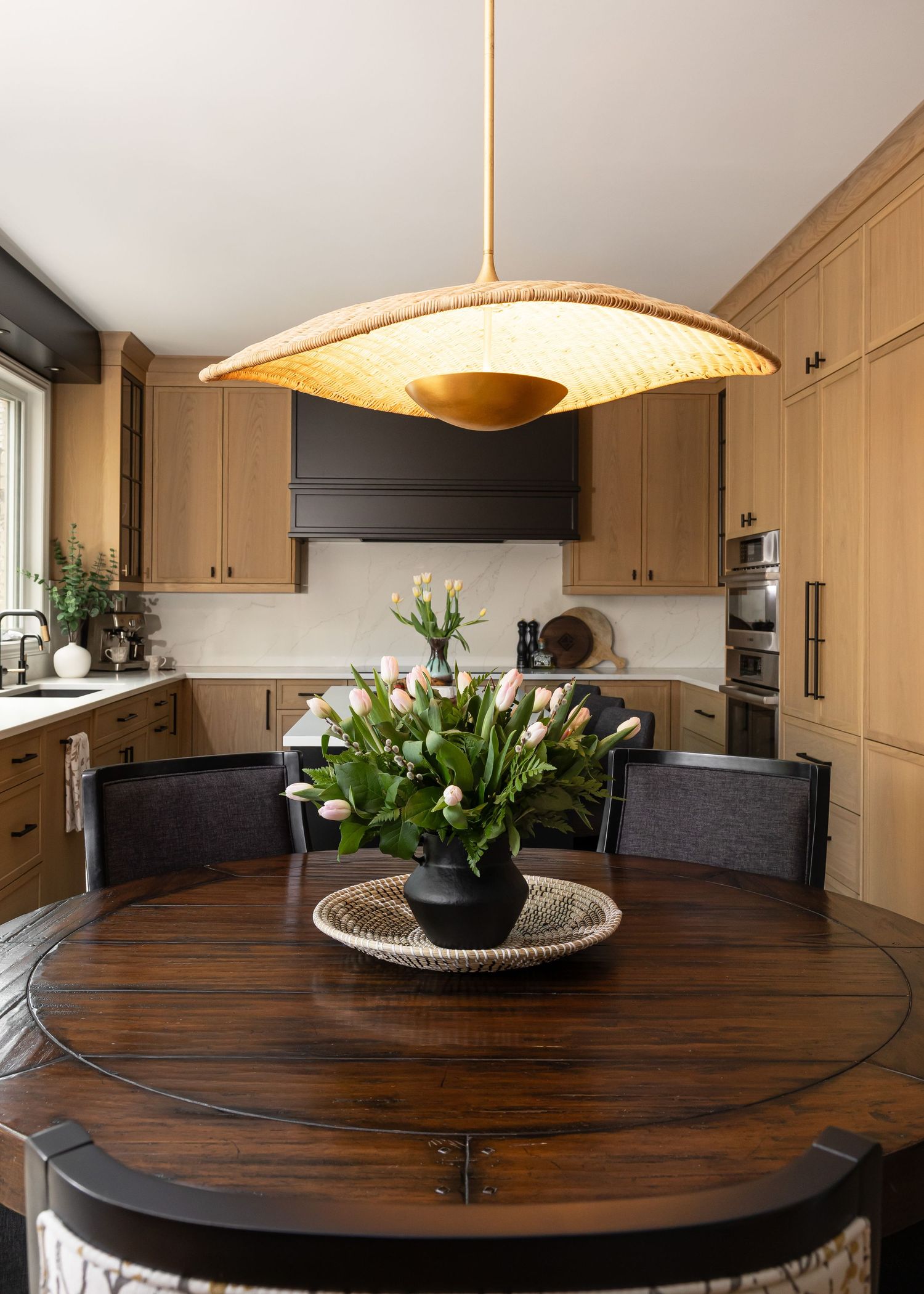 Modern kitchen with round wooden table, woven pendant light, and white floral centerpiece.