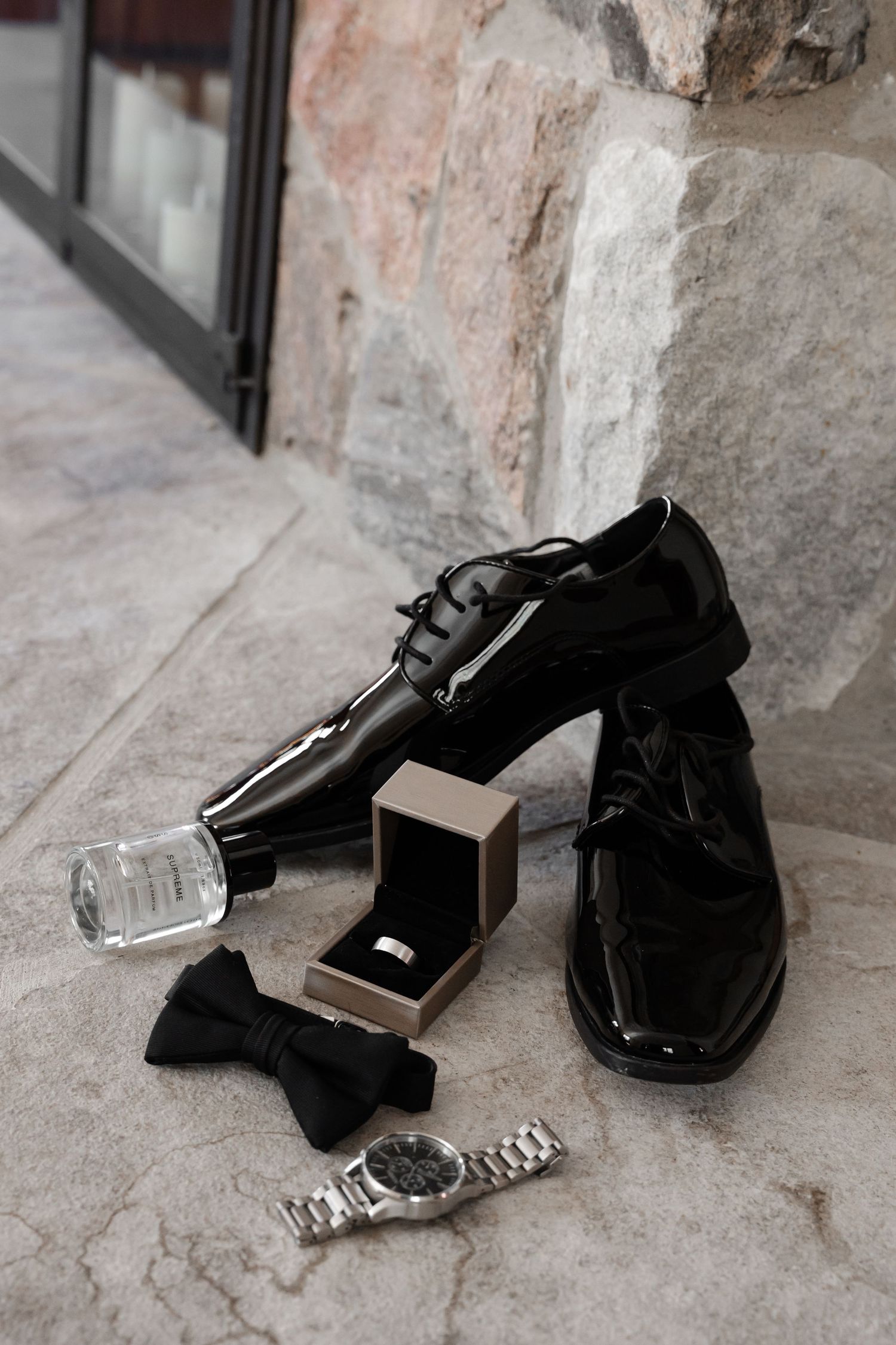 Groom's formal accessories including black patent shoes, bow tie and watch on stone floor.