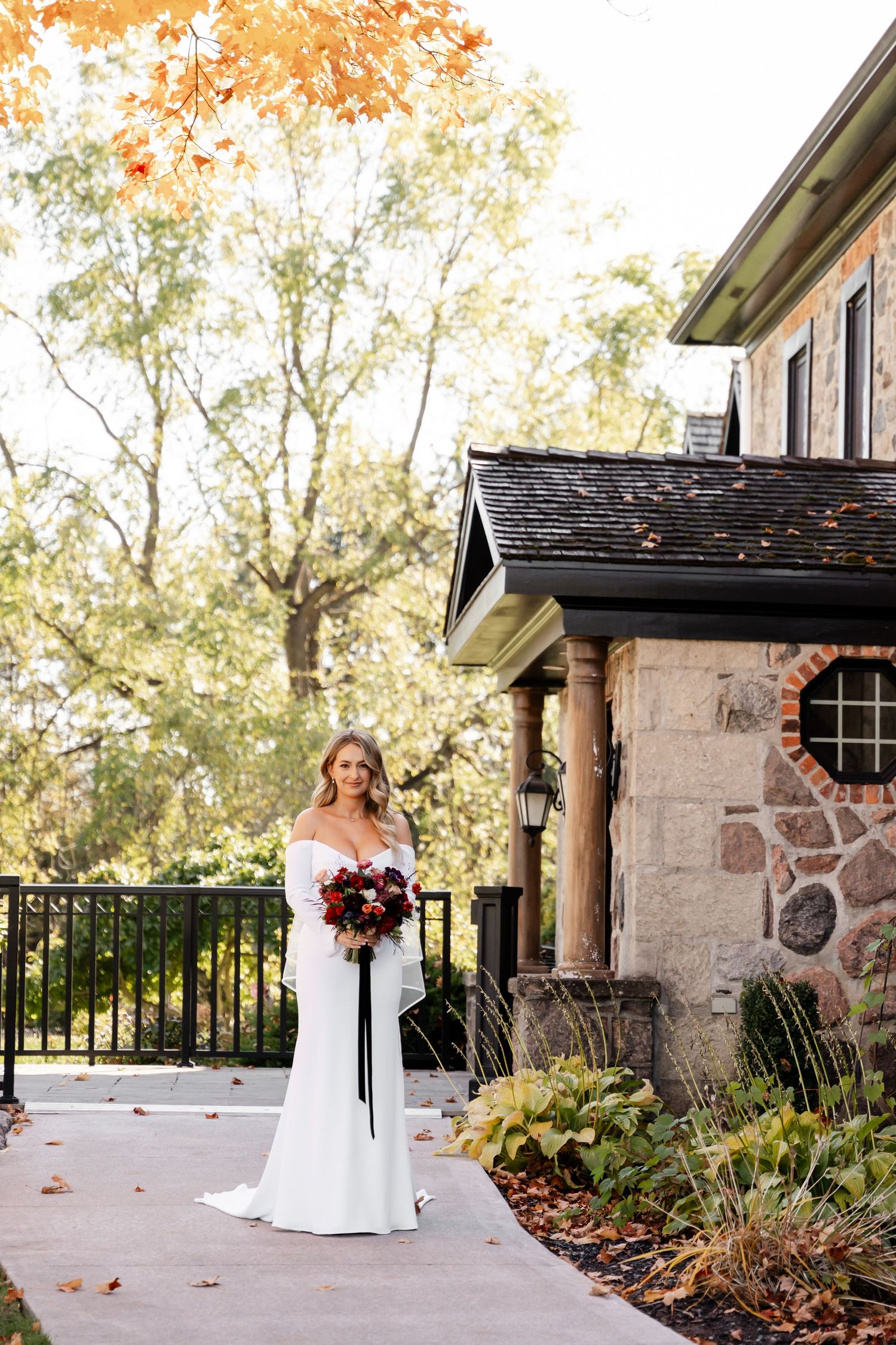 A bride stands elegantly in her white gown against an autumn stone building backdrop.