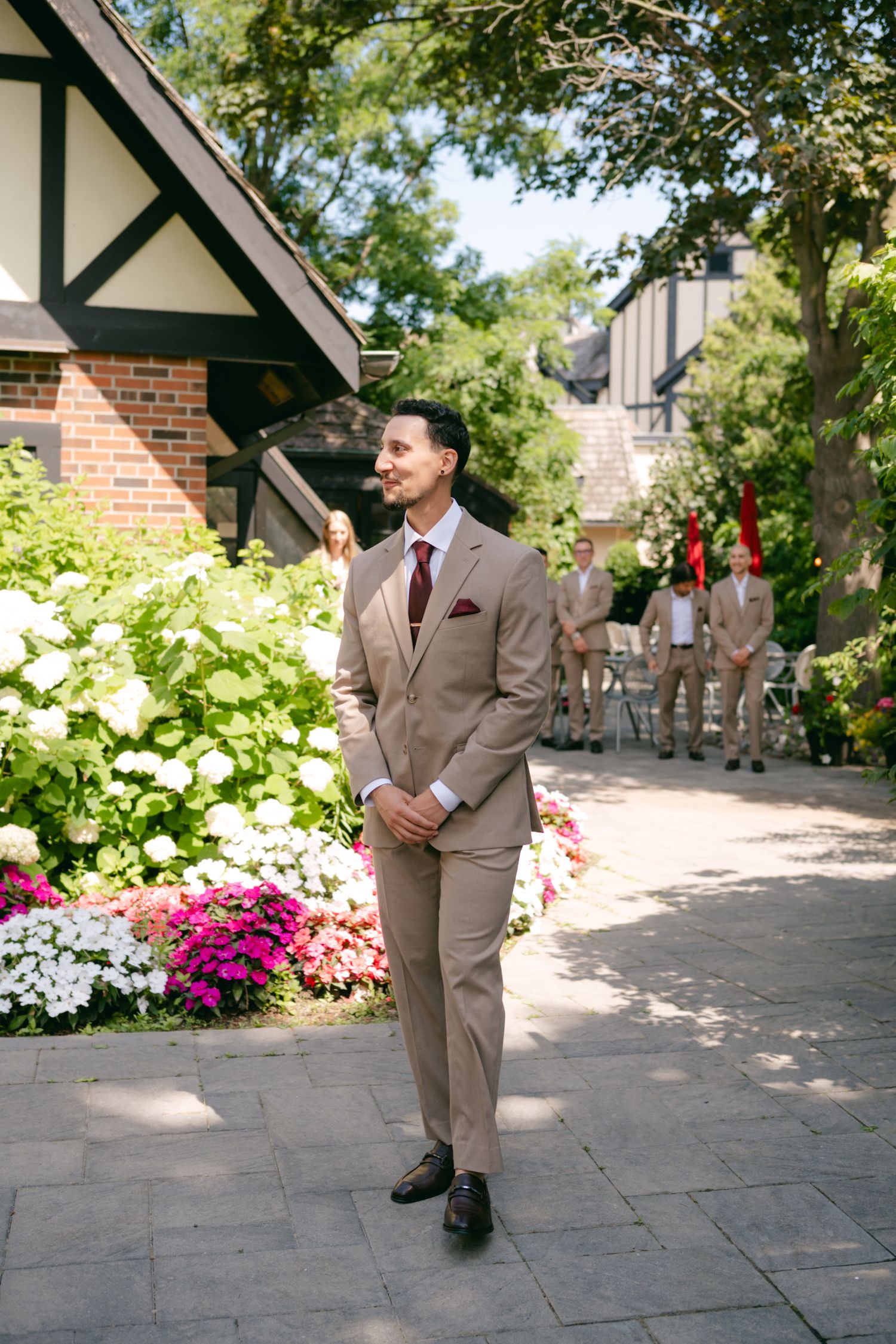 A groom in a tan suit standing on a stone pathway surrounded by colorful garden flowers.