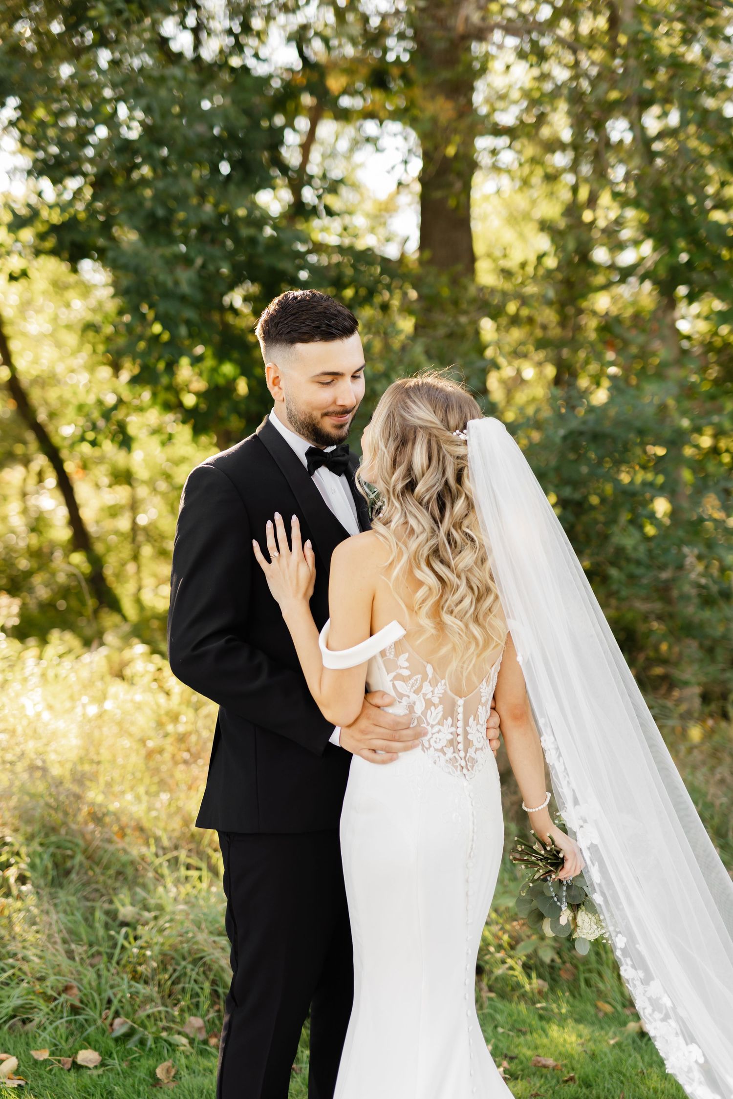 Elegant wedding couple embraces outdoors with detailed lace dress and long veil.