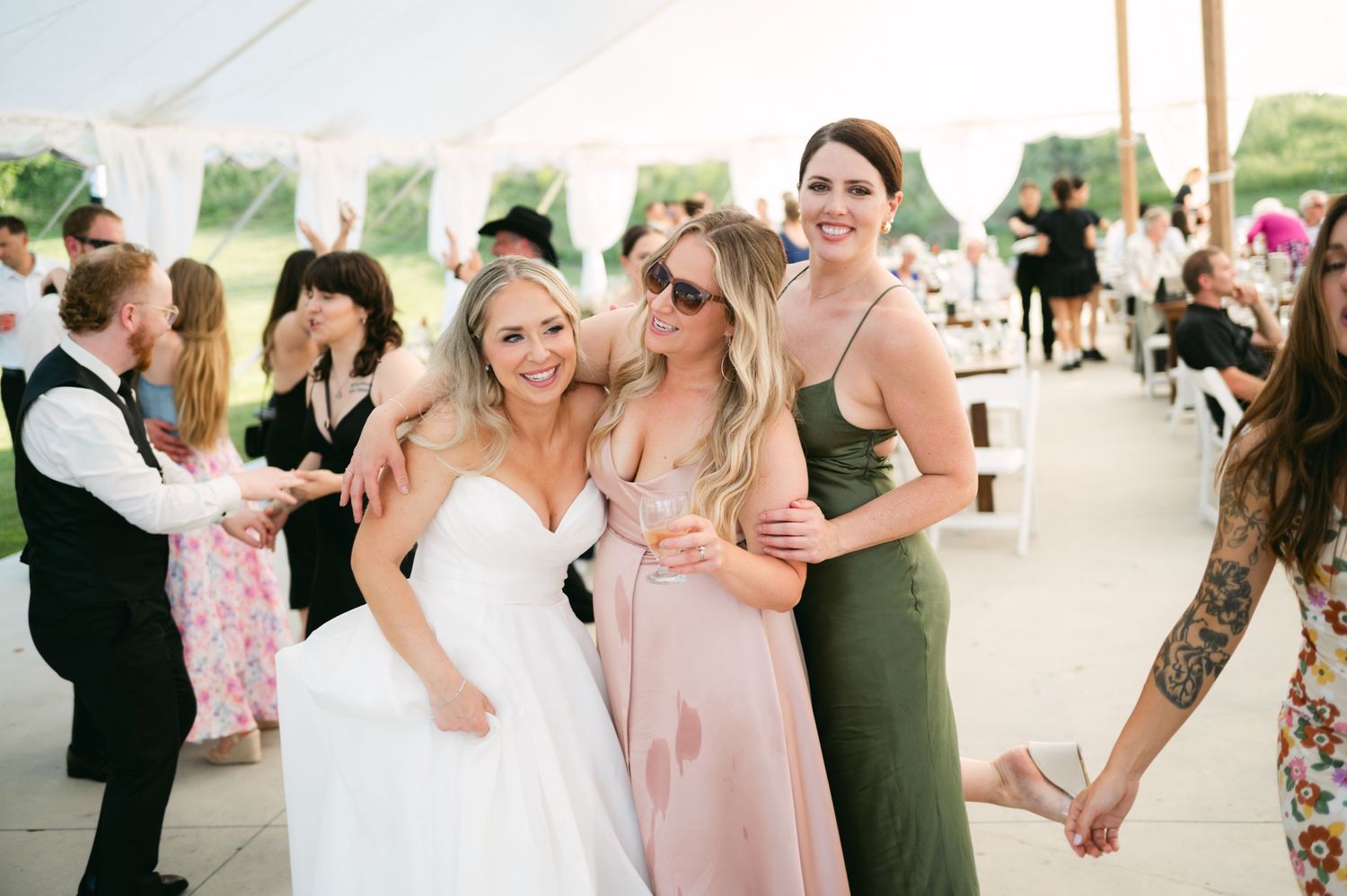 Wedding party members gather under white drapes wearing pink and floral dresses for group photos.