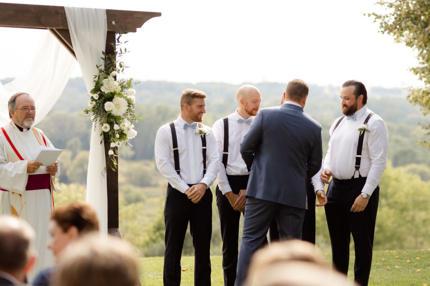 Wedding ceremony setup with officiant and groomsmen under wooden arch outdoors.