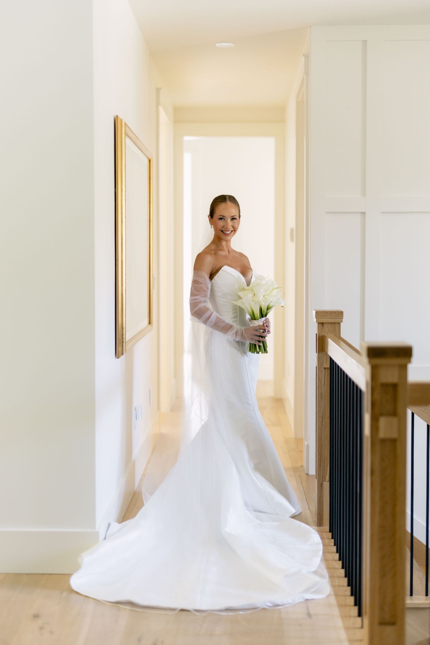 Radiant bride in strapless mermaid gown holding white calla lily bouquet in hallway.