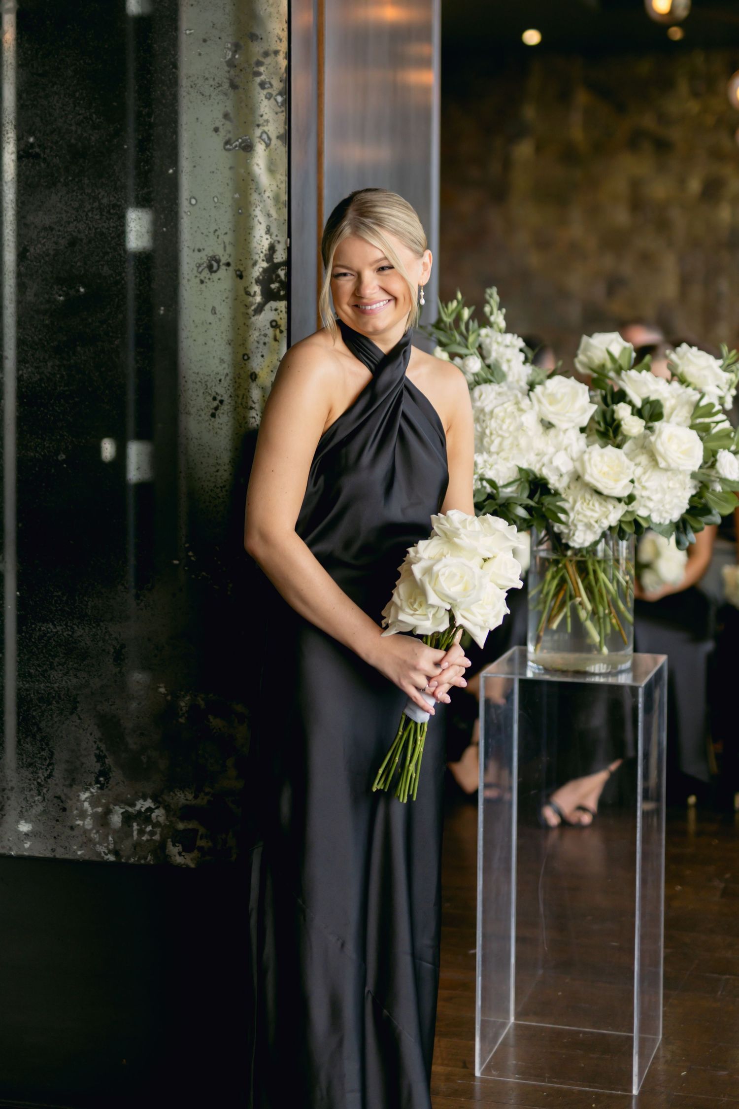 Bridesmaid in black halter gown smiles holding white rose bouquet beside floral arrangement on acrylic stand.