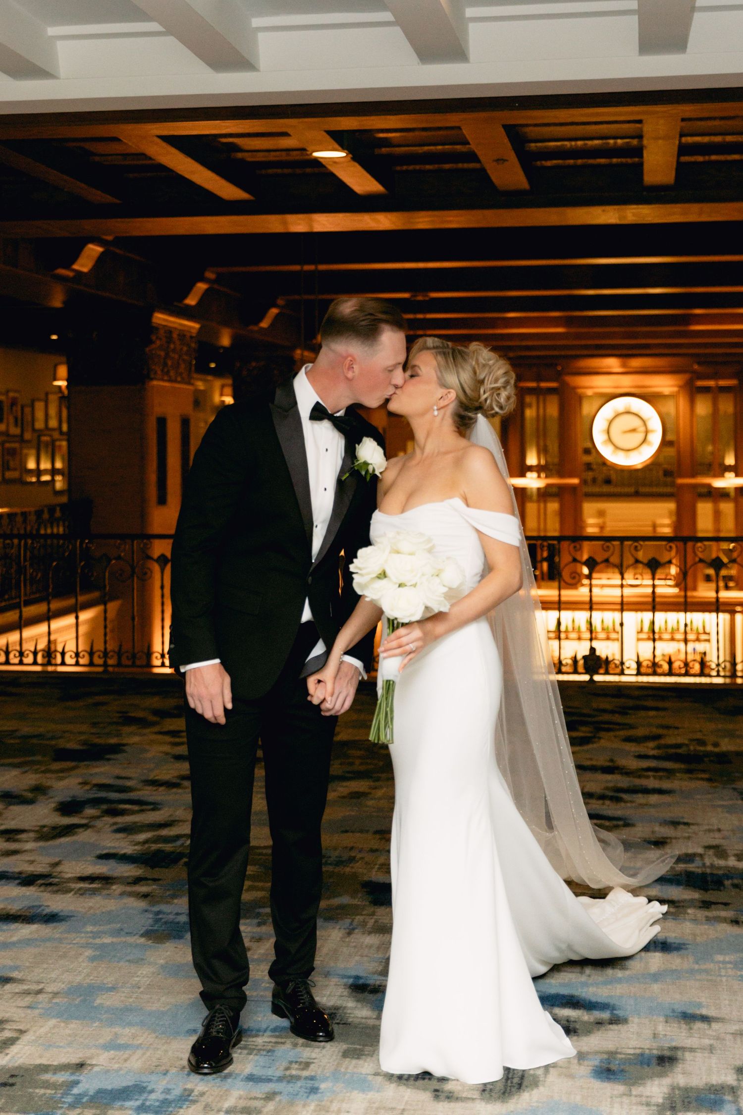 Bride and groom share a kiss in an elegant hotel lobby, bride holding white bouquet in fitted off-shoulder gown.
