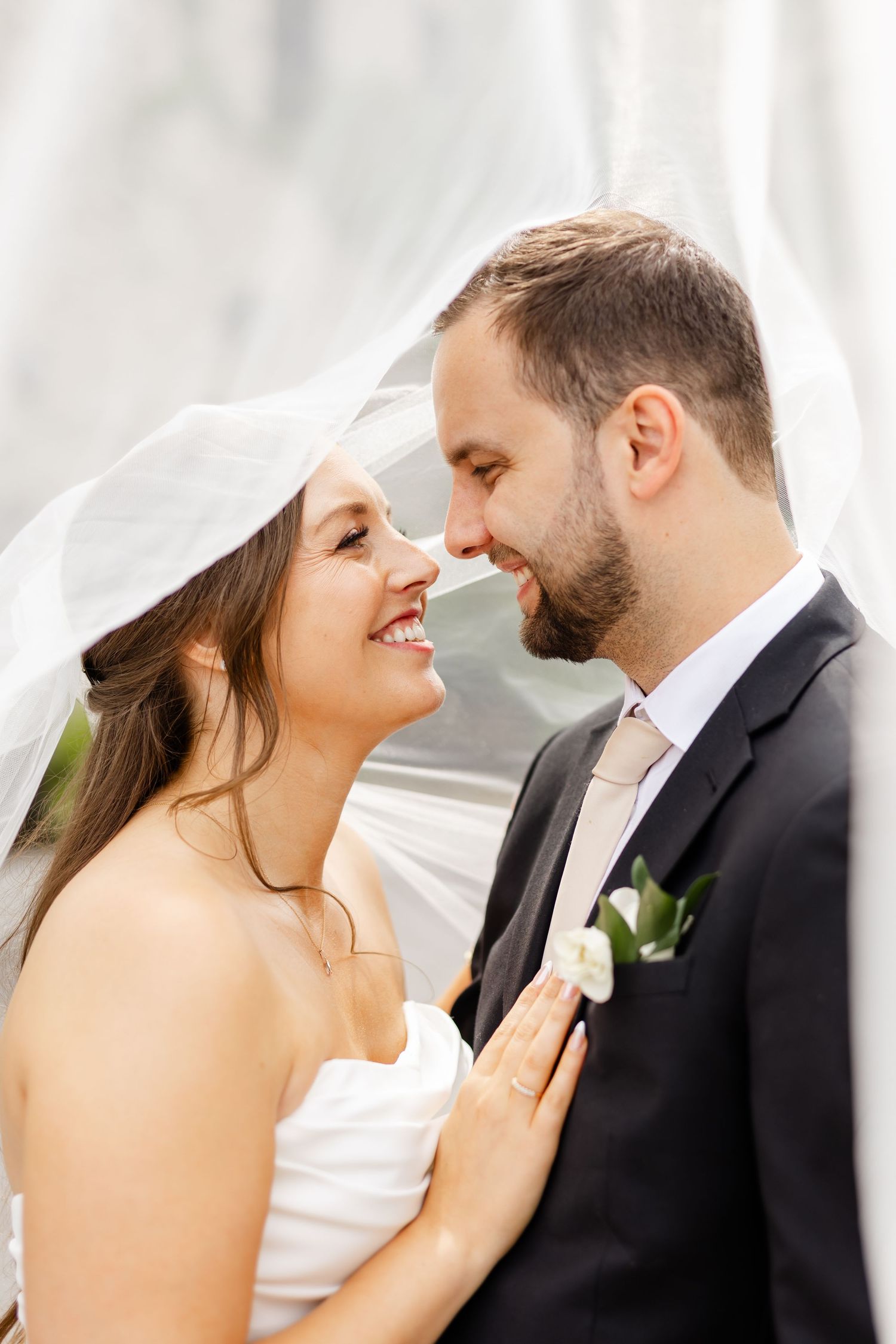 Bride and groom share romantic moment under flowing veil, smiling and touching noses on wedding day.