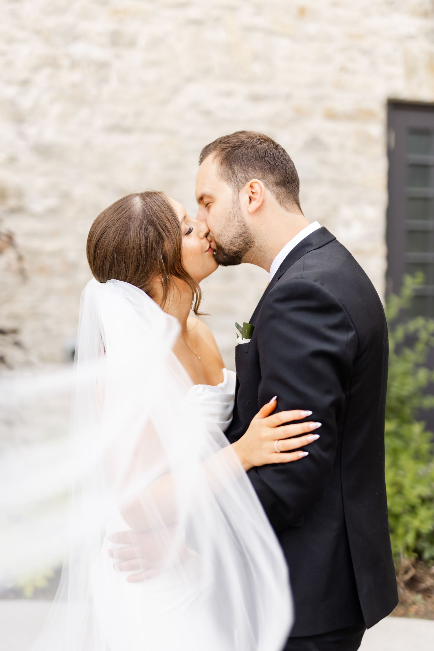 Bride and groom share a romantic kiss outdoors with veil flowing in front of stone building.