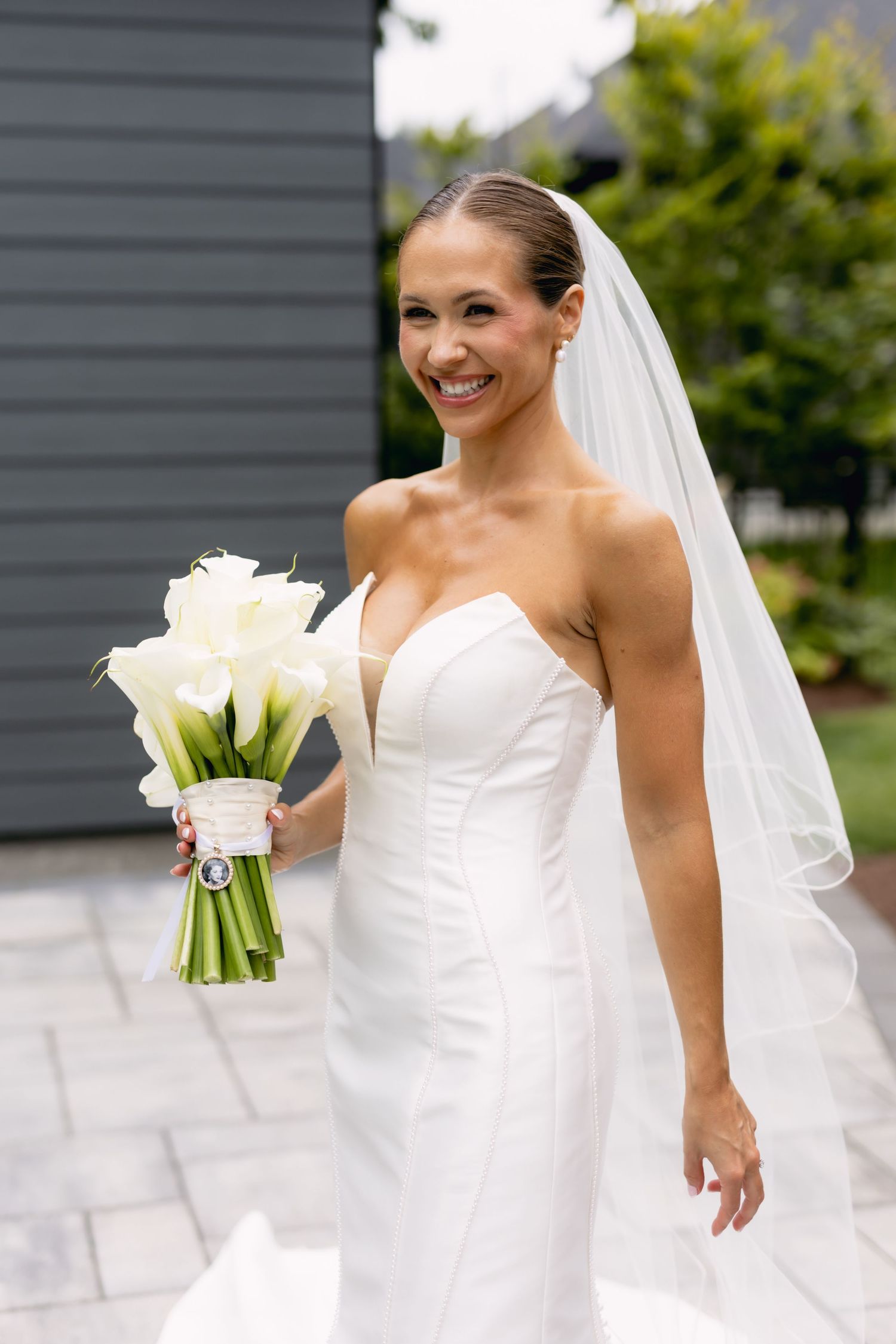 Smiling bride in strapless white gown and veil holds white calla lily bouquet outdoors on stone patio.