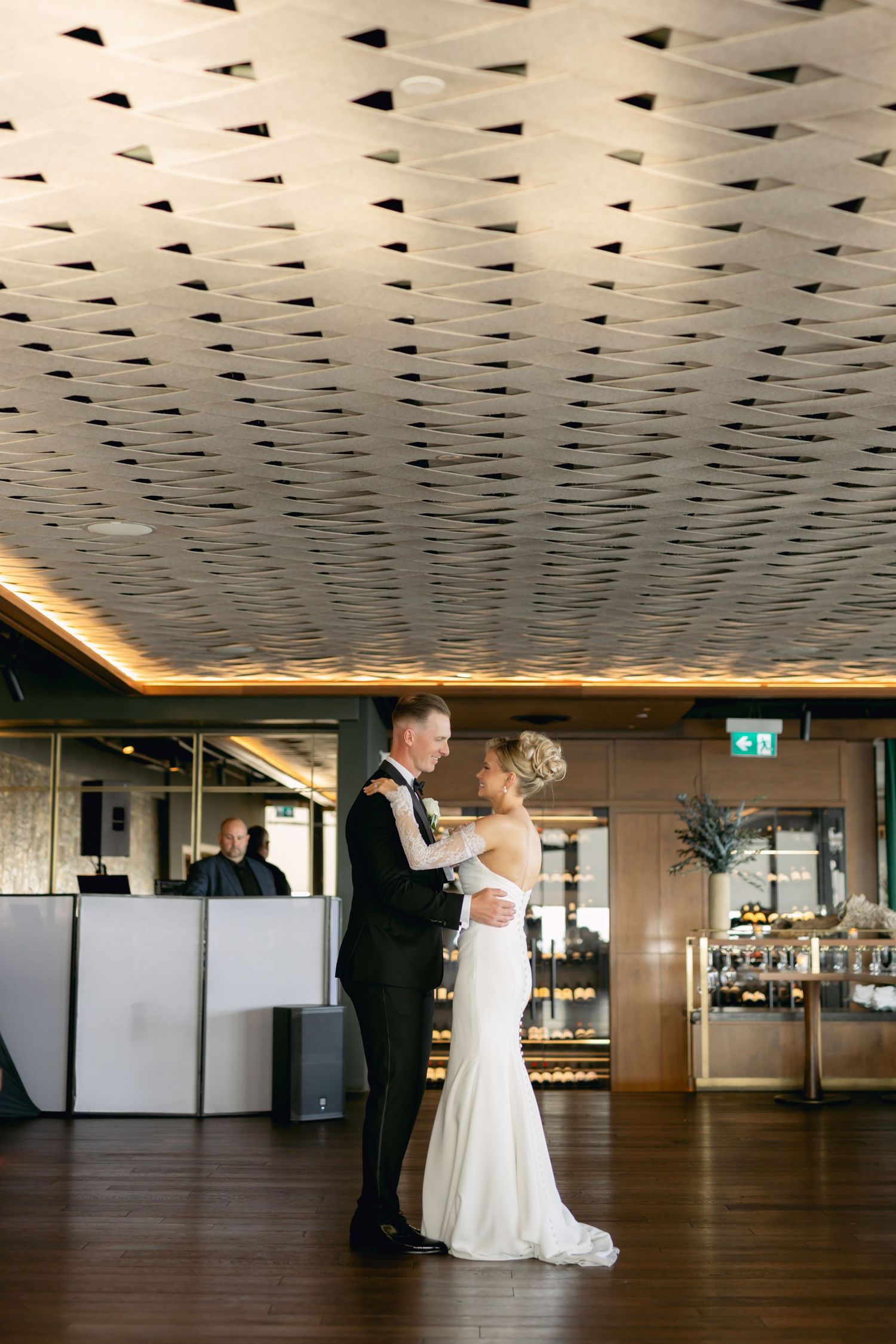 Bride and groom share first dance in elegant venue with geometric ceiling and warm ambient lighting.