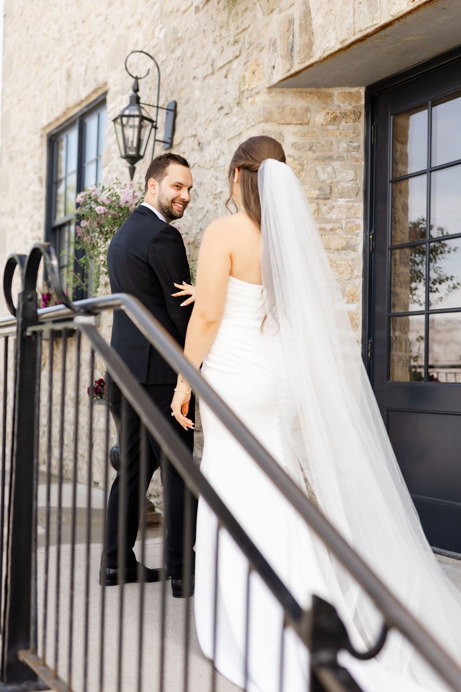Bride and groom share a romantic moment on stone steps outside a rustic venue, with her white veil trailing behind.