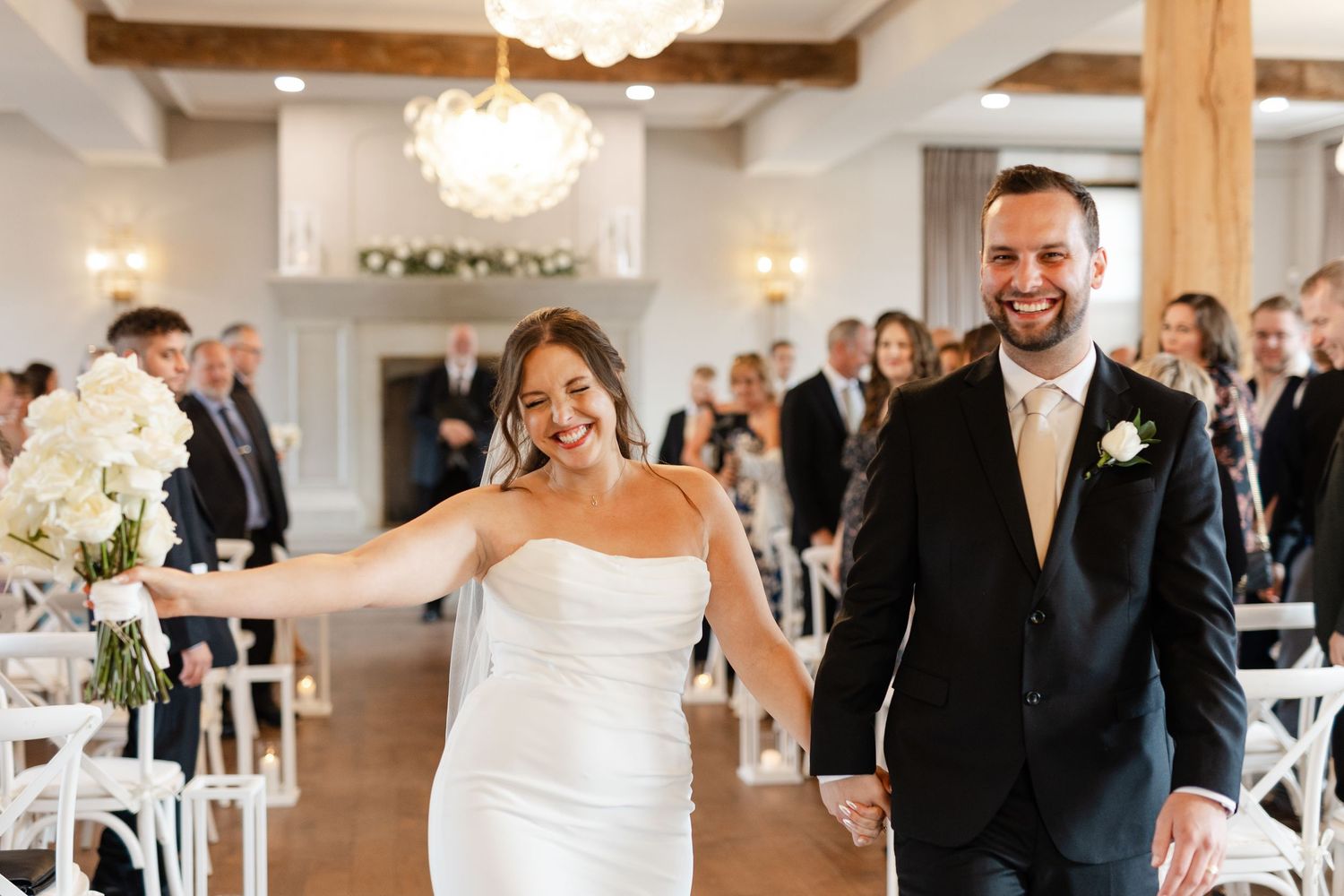 Newlyweds smile and hold hands while walking back down the aisle after their indoor wedding ceremony.