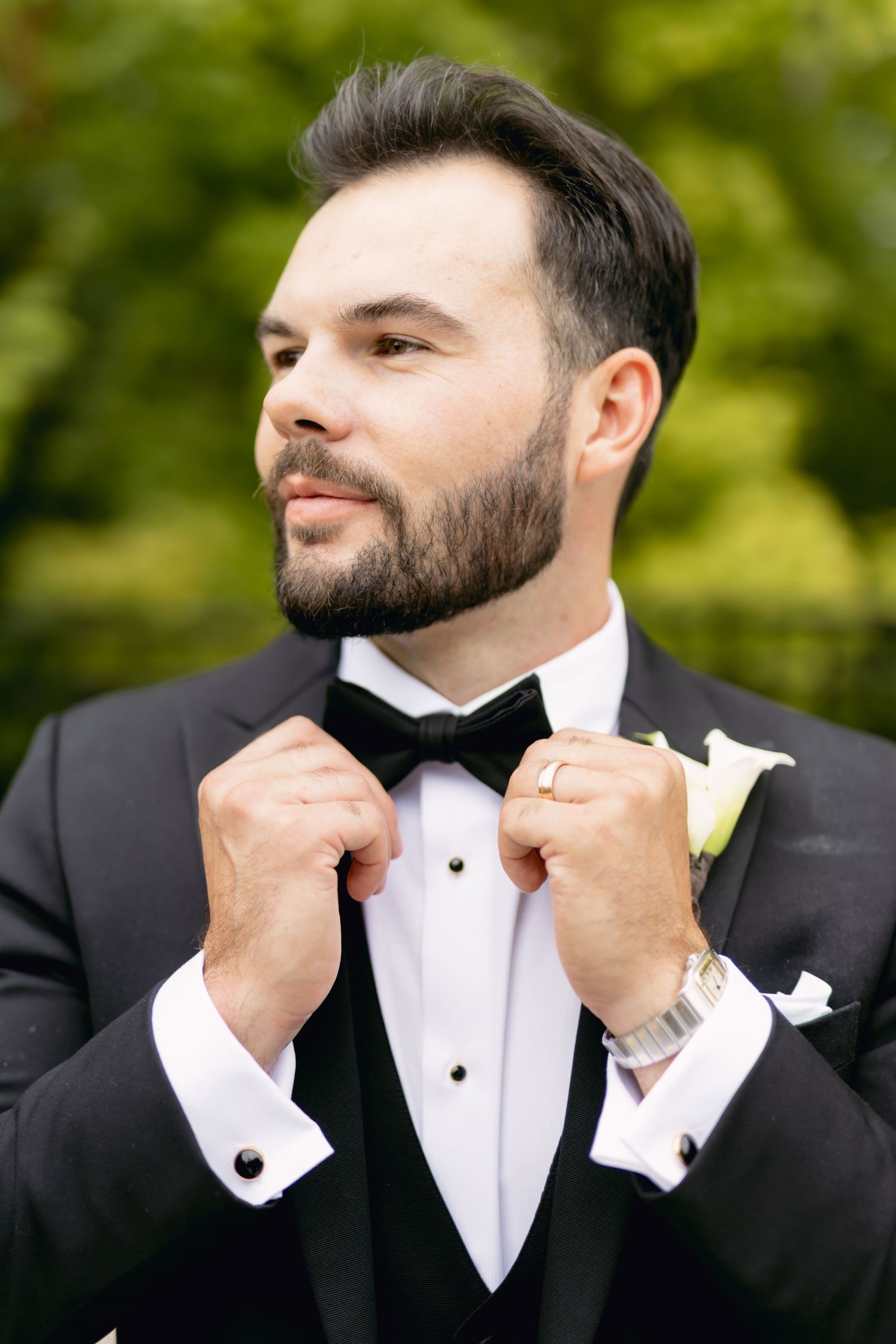 Dapper groom in black tuxedo adjusting bow tie with white calla lily boutonniere outdoors.