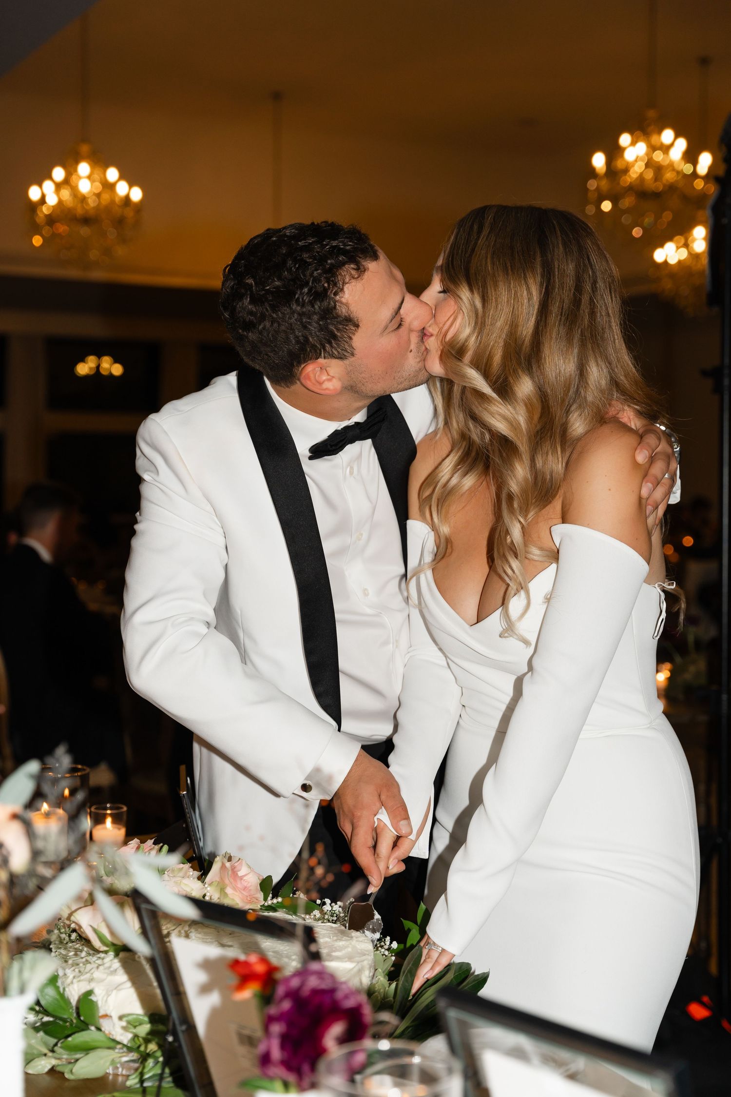 Couple shares romantic kisses during wedding reception with elegant chandeliers in the background.