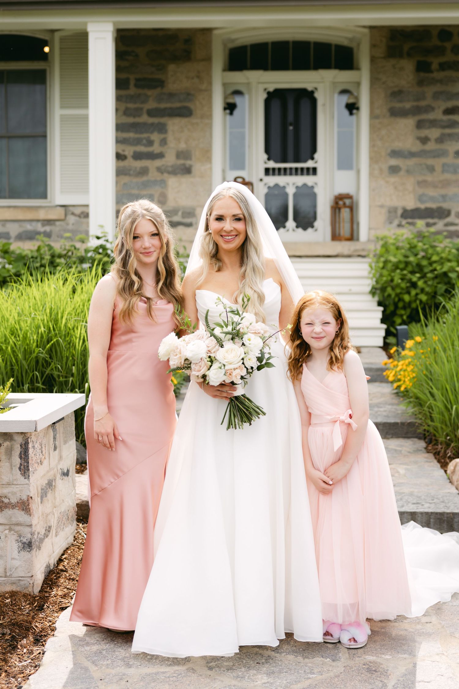 Wedding party members in pink and white dresses pose together on stone steps.