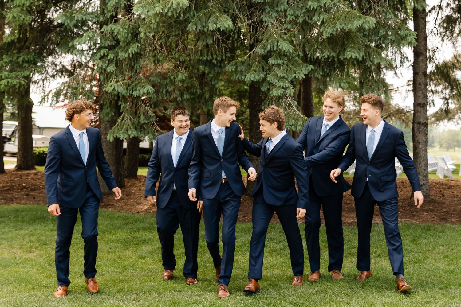 Wedding party members in navy suits leap joyfully across grass during outdoor photo session.