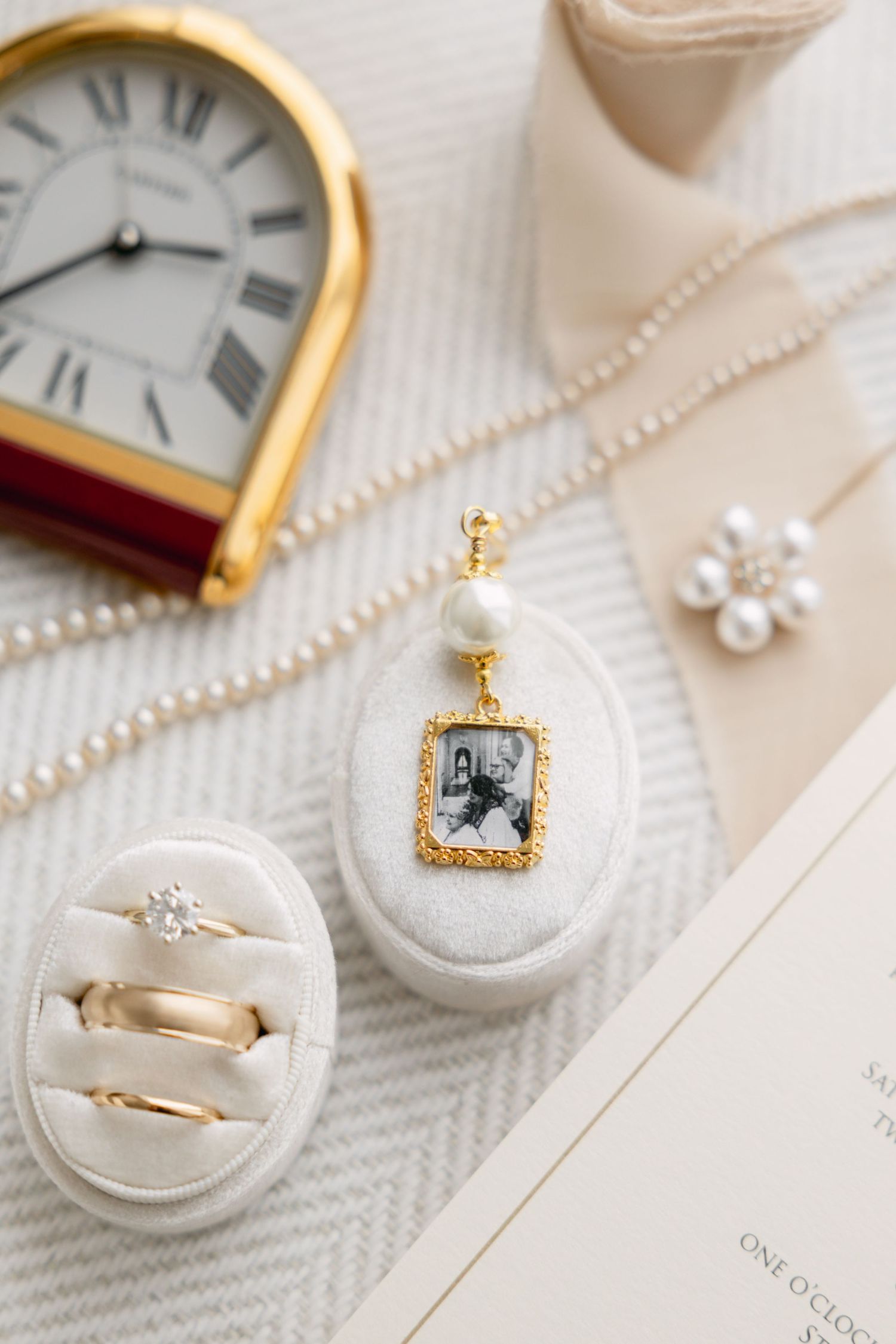Gold photo locket pendant with pearl, wedding rings in velvet box, pearl necklace, and vintage clock styled flat lay.