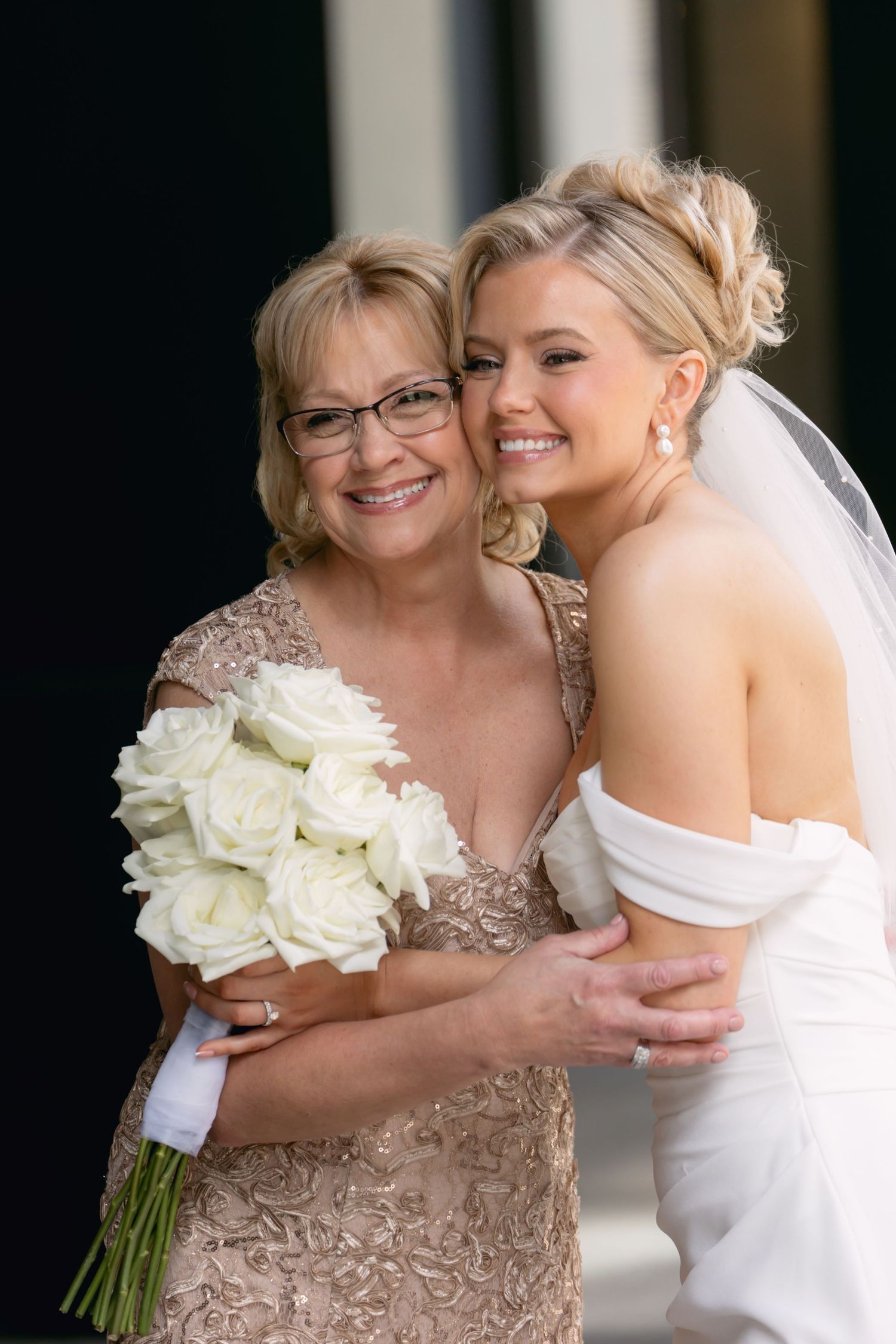 Bride in white off-shoulder gown embraces smiling mother in gold lace dress holding white rose bouquet.