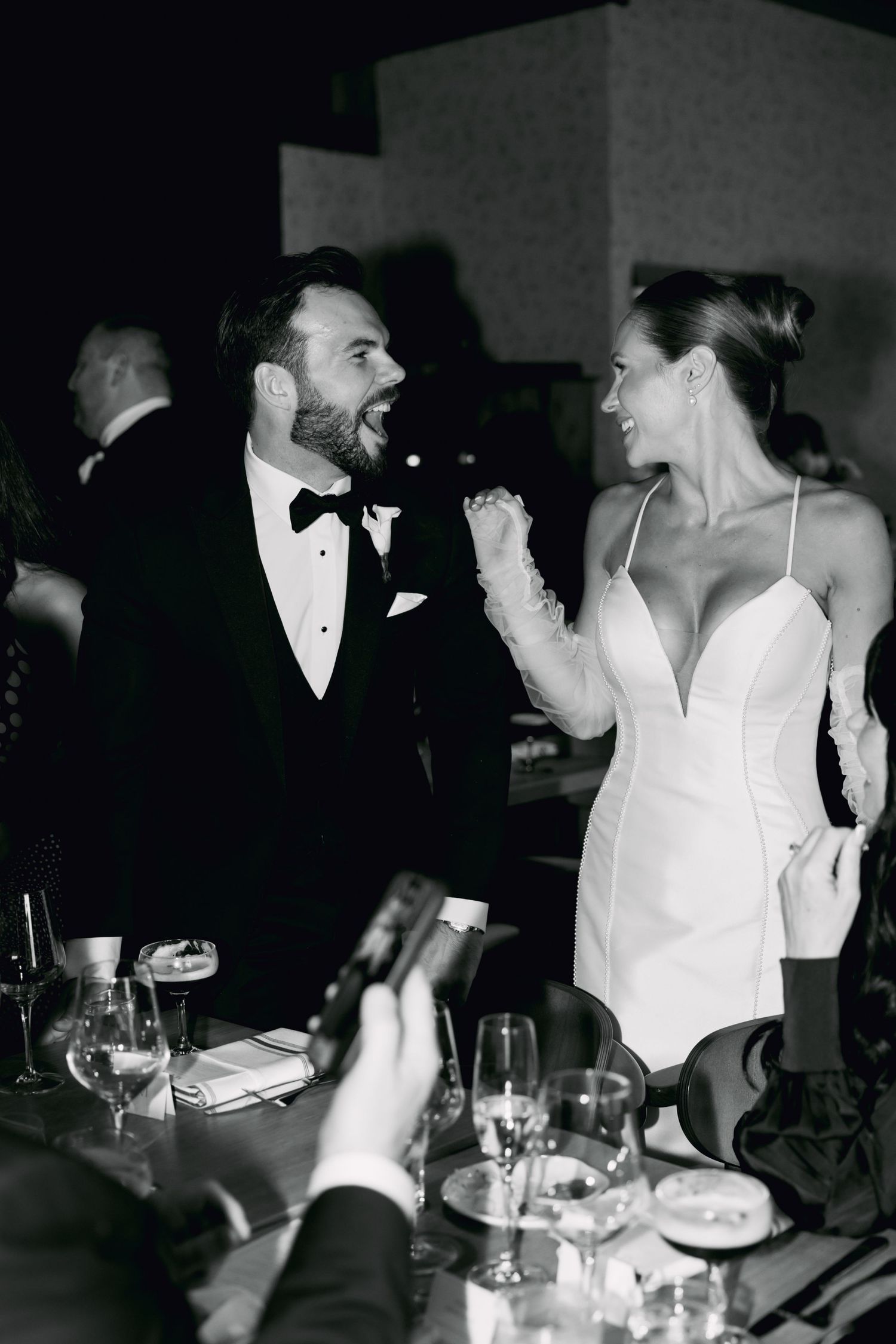 Black and white photo of bride in white mini dress and groom in tuxedo laughing at reception.