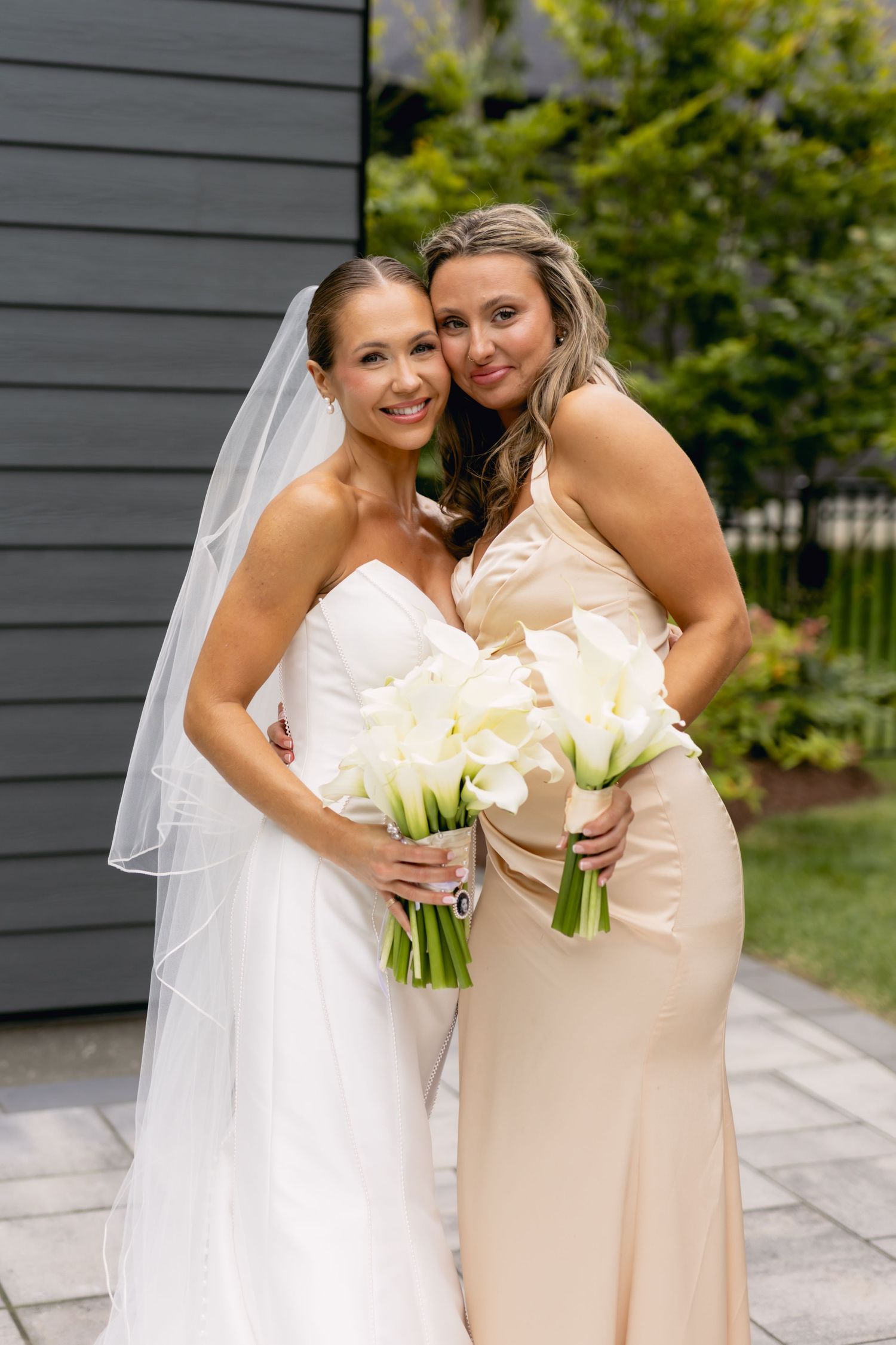 Bride in strapless white gown and veil poses with bridesmaid in champagne dress holding white calla lily bouquets.