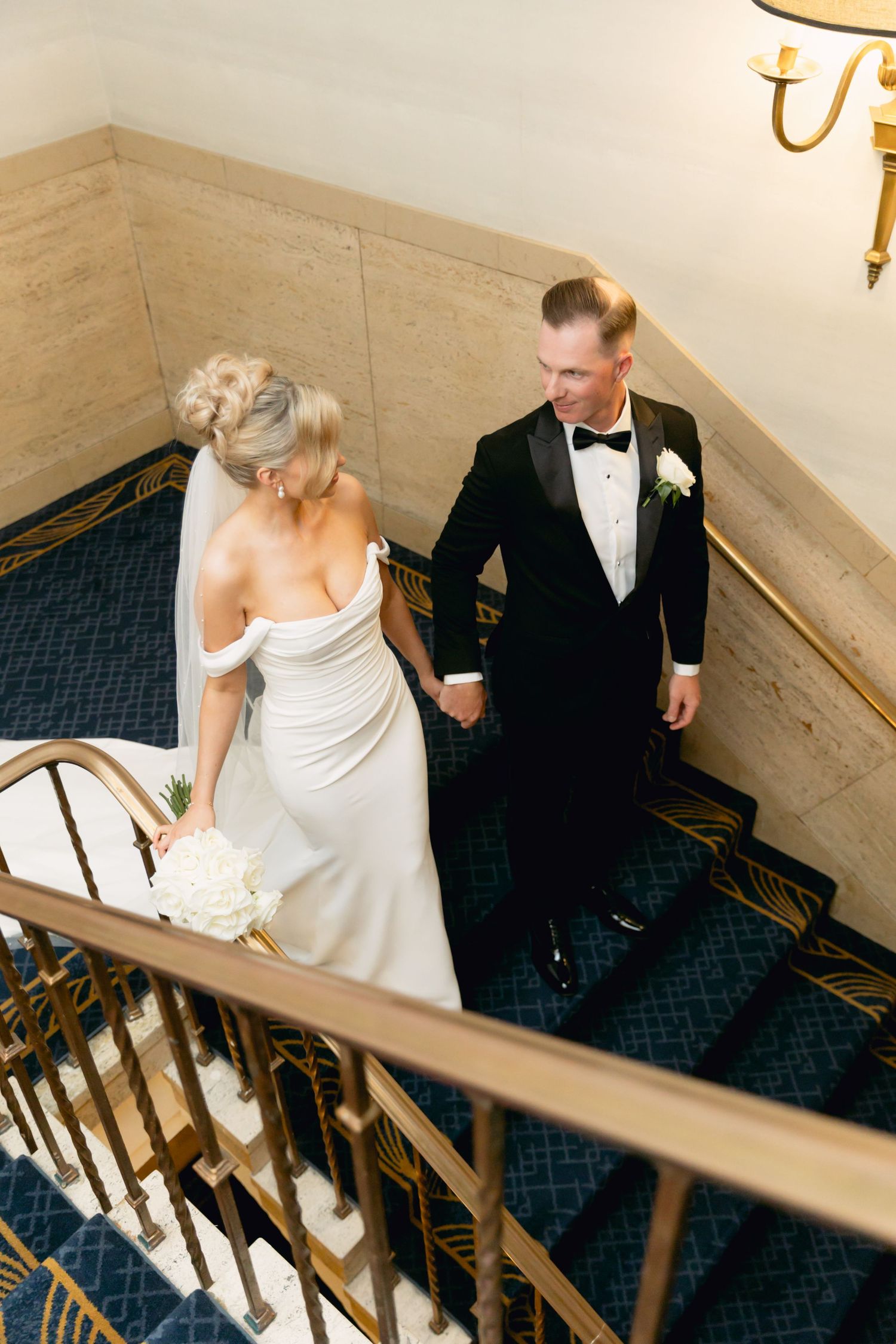 Bride and groom holding hands walking down elegant staircase, bride in off-shoulder white gown holding bouquet.