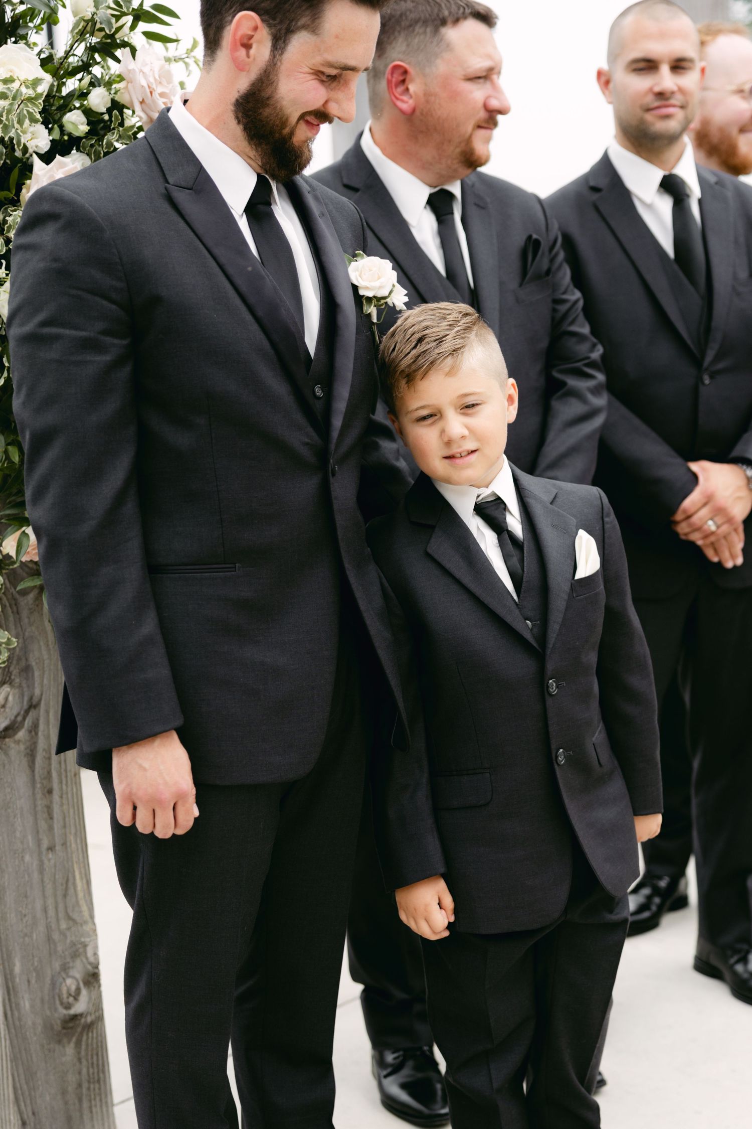 Groomsmen in black suits standing together at outdoor wedding ceremony.