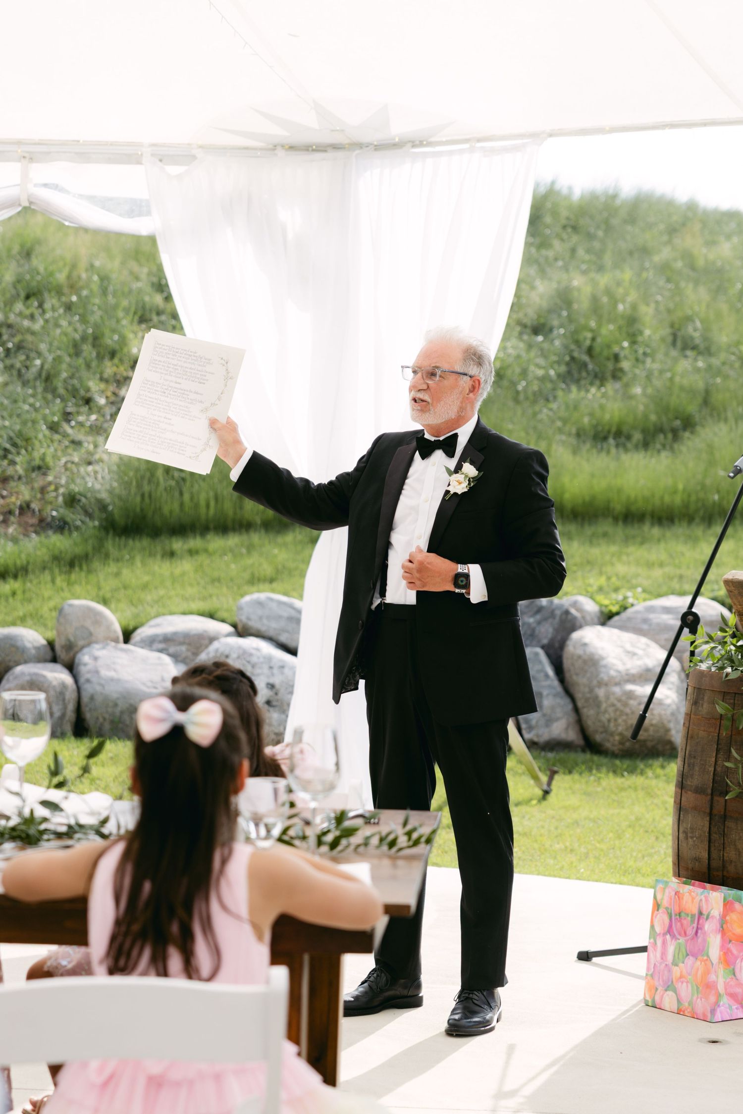 Wedding guests laugh during a playful moment at an outdoor reception under a white tent.