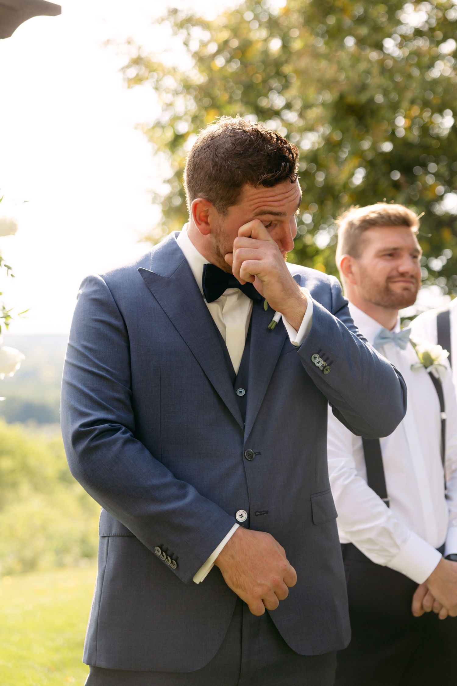 Groomsmen in navy suits share emotional reactions during outdoor wedding ceremony.