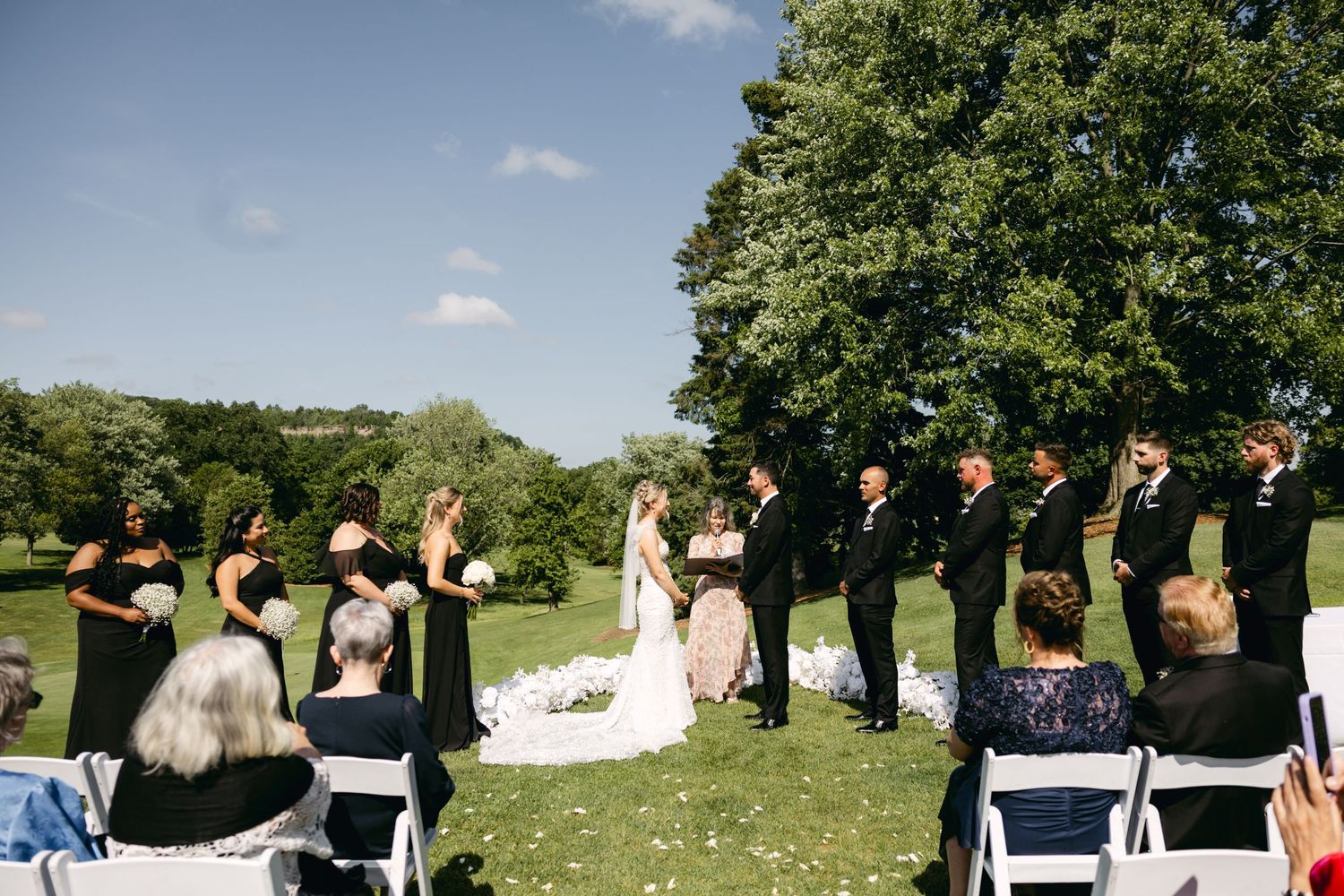 Wedding guests seated outdoors on white chairs during a sunny garden ceremony.