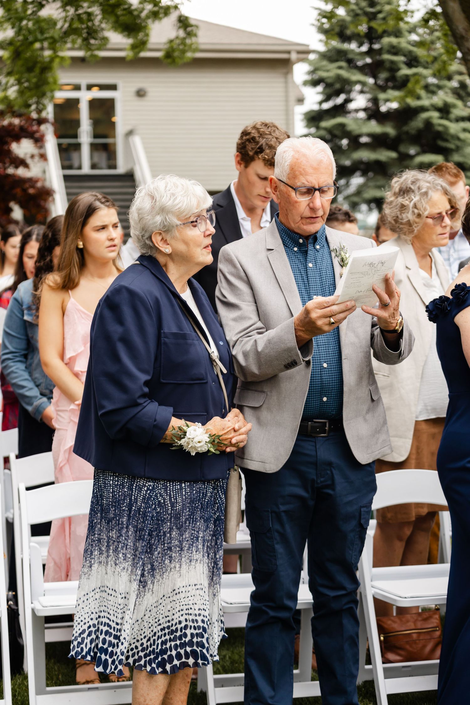 Wedding guests holding programs during outdoor ceremony with white house in background.
