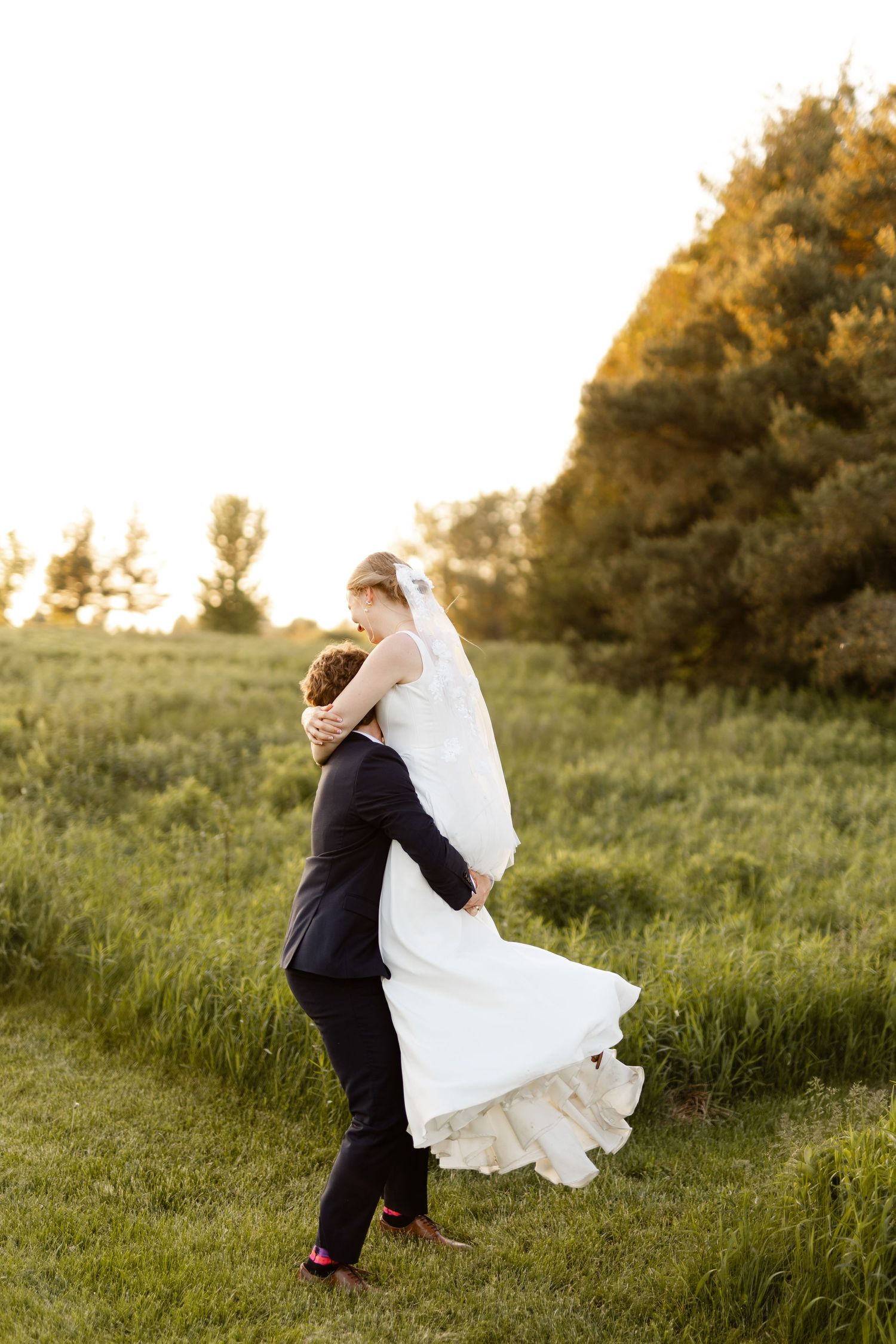 Playful wedding couple dancing and spinning in a grassy field at sunset.