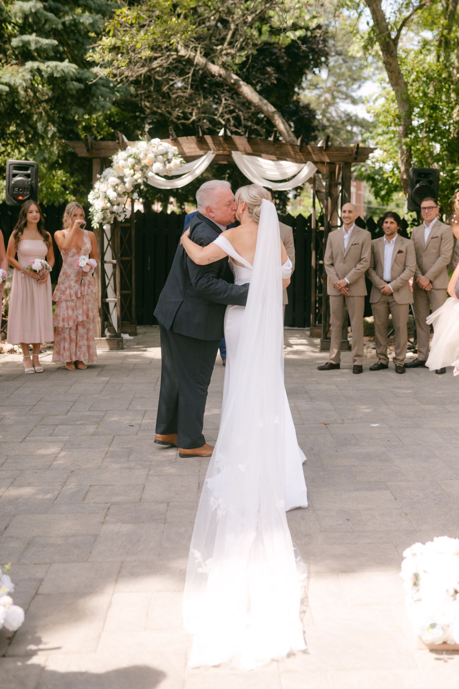 Wedding ceremony setup with white decorations and guests in formal attire on a stone patio.