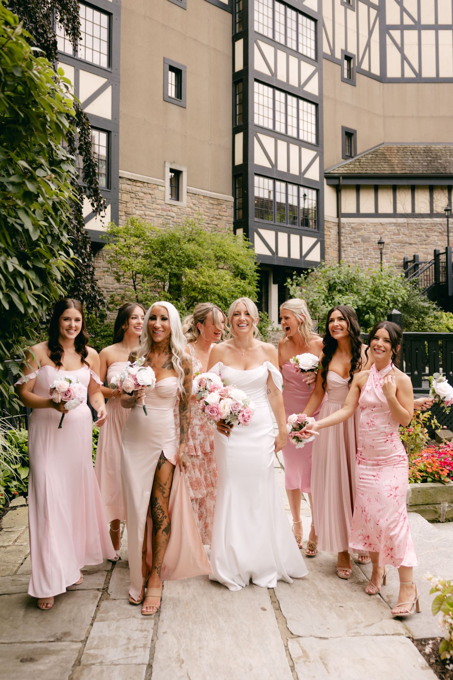 Wedding party in pink dresses poses outdoors against stone building backdrop.