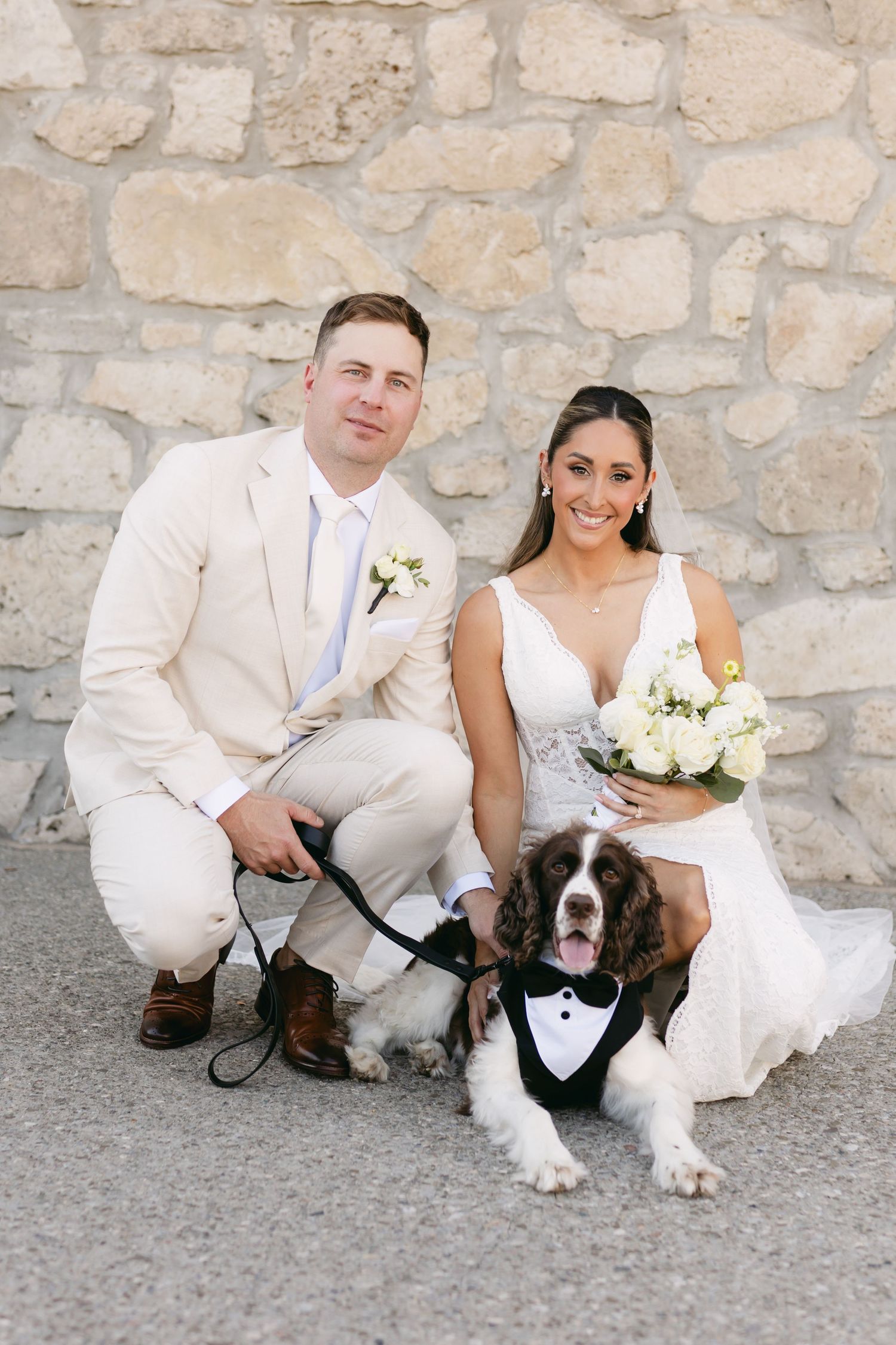 Newlyweds in white attire kneel beside their dressed up dog for wedding photos.