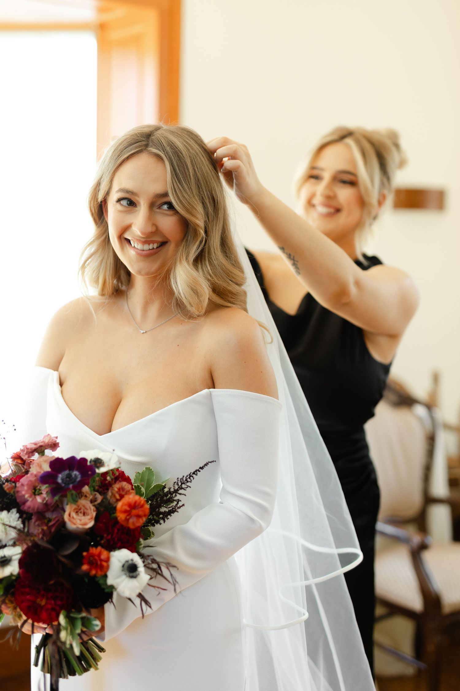 Bride in white off-shoulder dress holding vibrant burgundy and pink floral bouquet during wedding preparations.