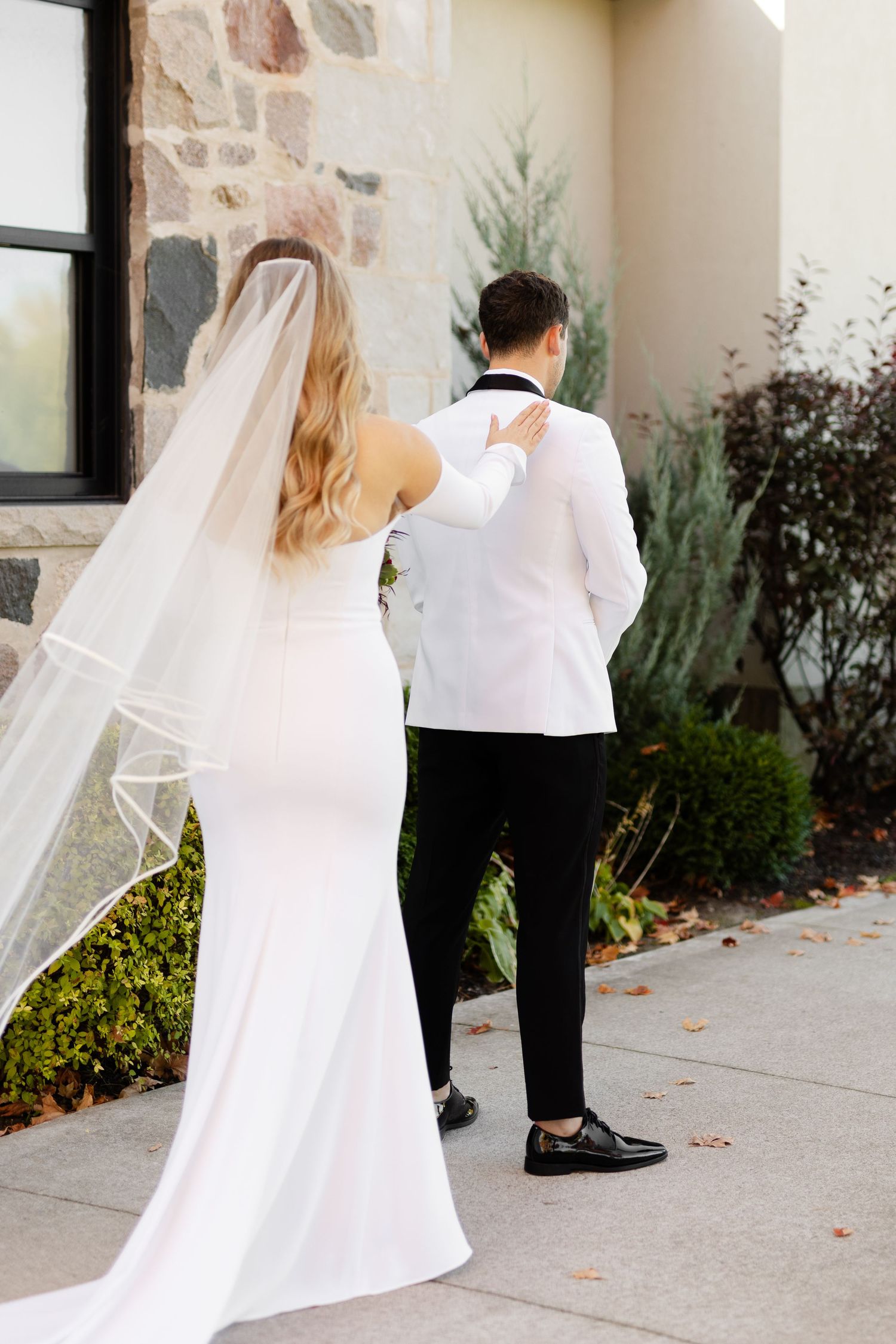 A bride in a sleek white gown approaches her groom from behind during their first look.
