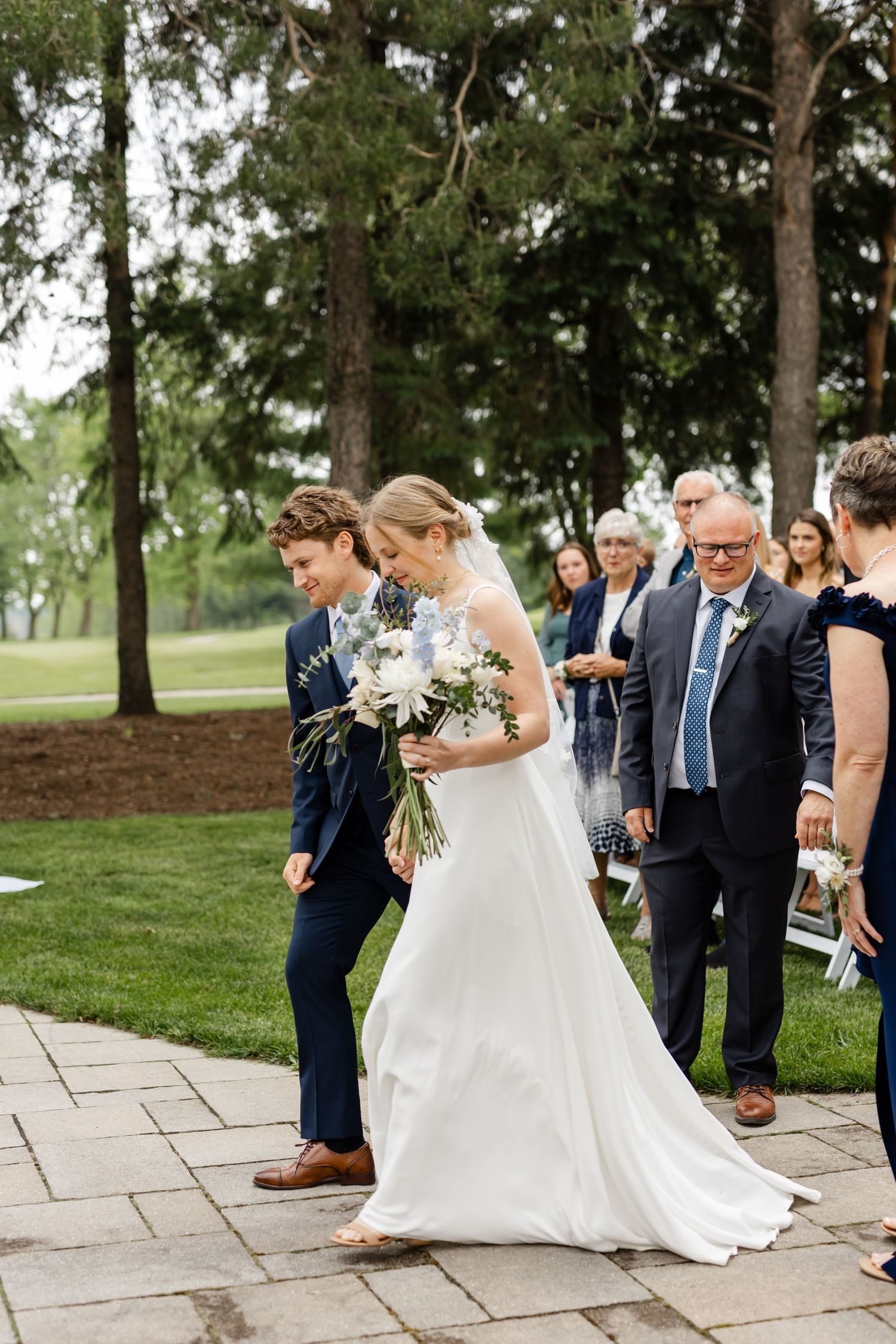 Wedding party walking along stone path outdoors with bride in white dress holding bouquet.