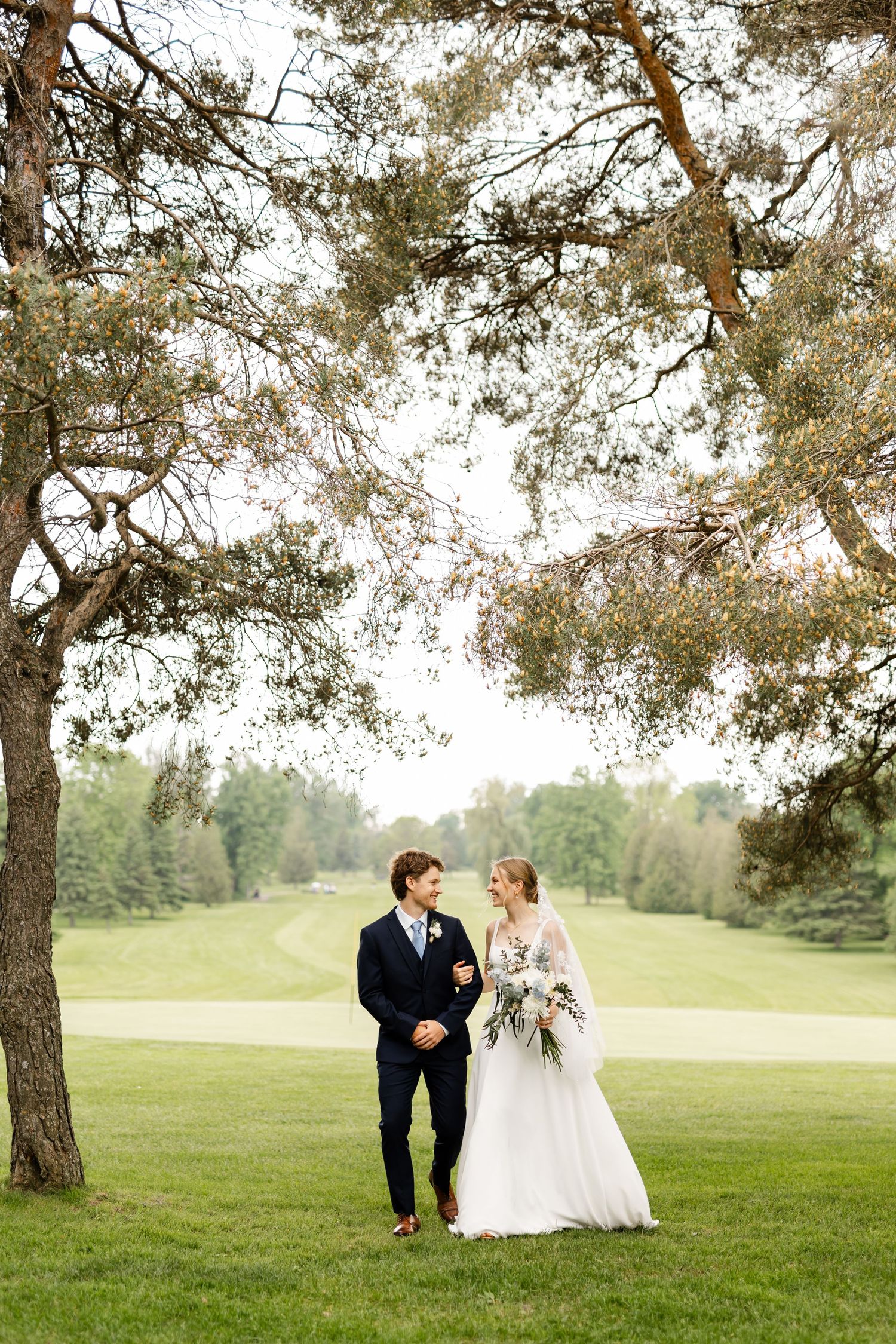 A couple in wedding attire walks together on a grassy hillside under tall pine trees.