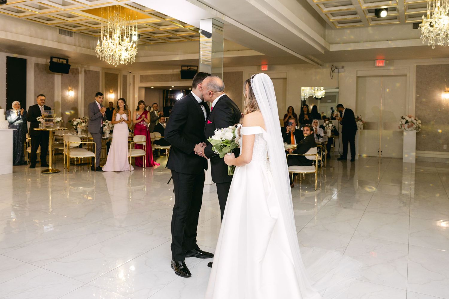 A couple shares a moment during their wedding ceremony in an elegant white ballroom.