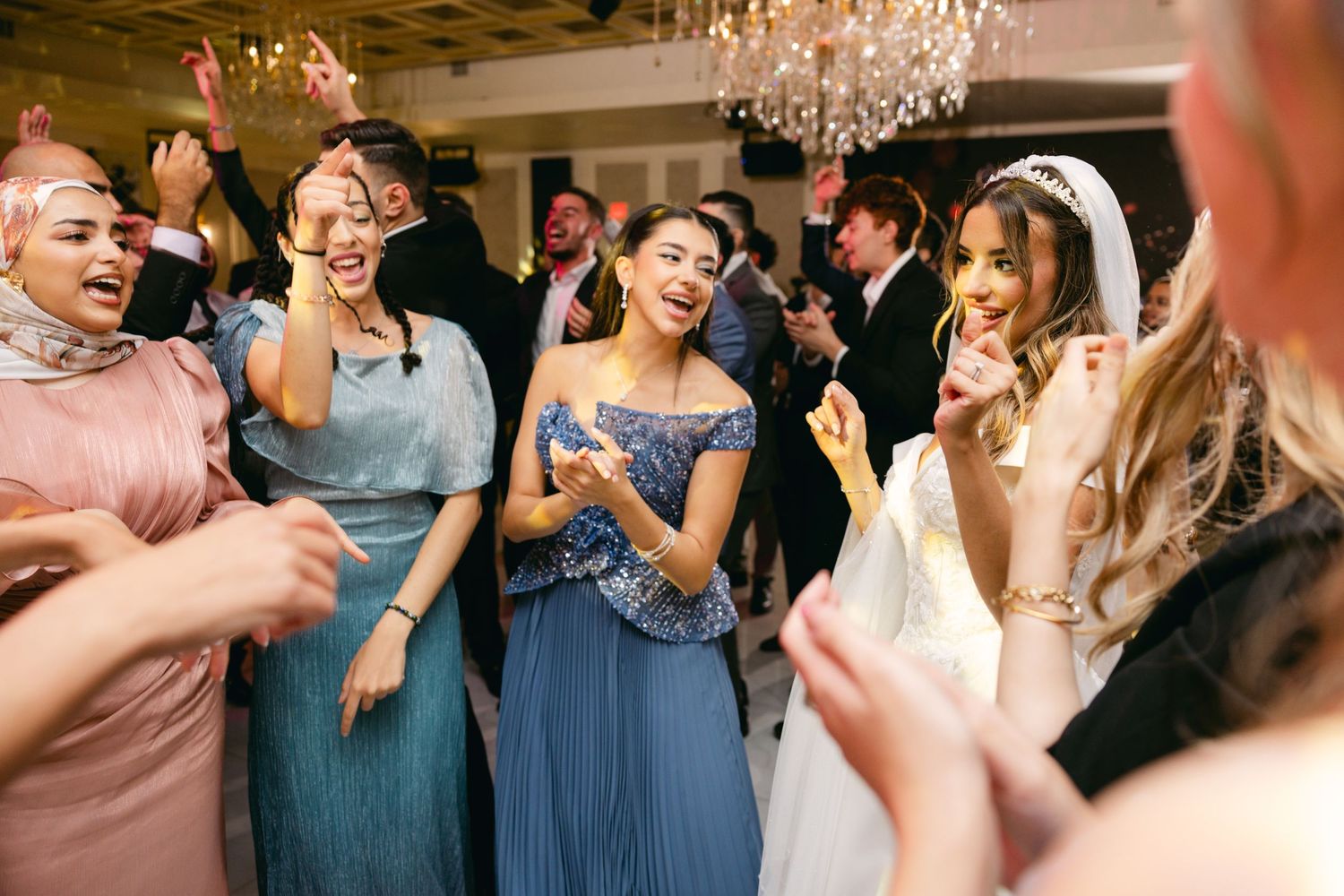 Wedding guests in colorful dresses laugh and dance together under elegant crystal chandeliers at a reception.