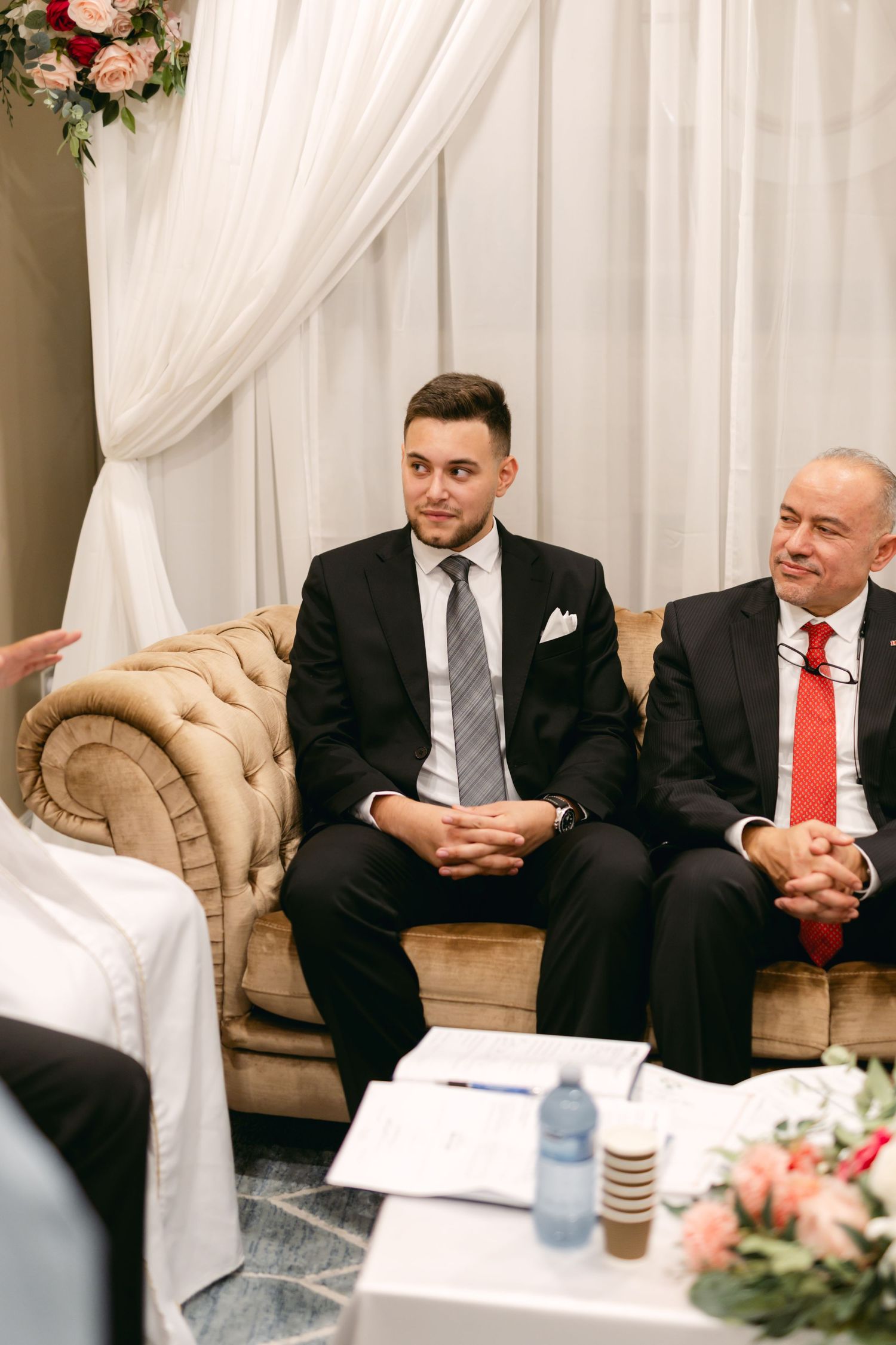 People seated on beige tufted chairs during a formal meeting with white drape backdrop.