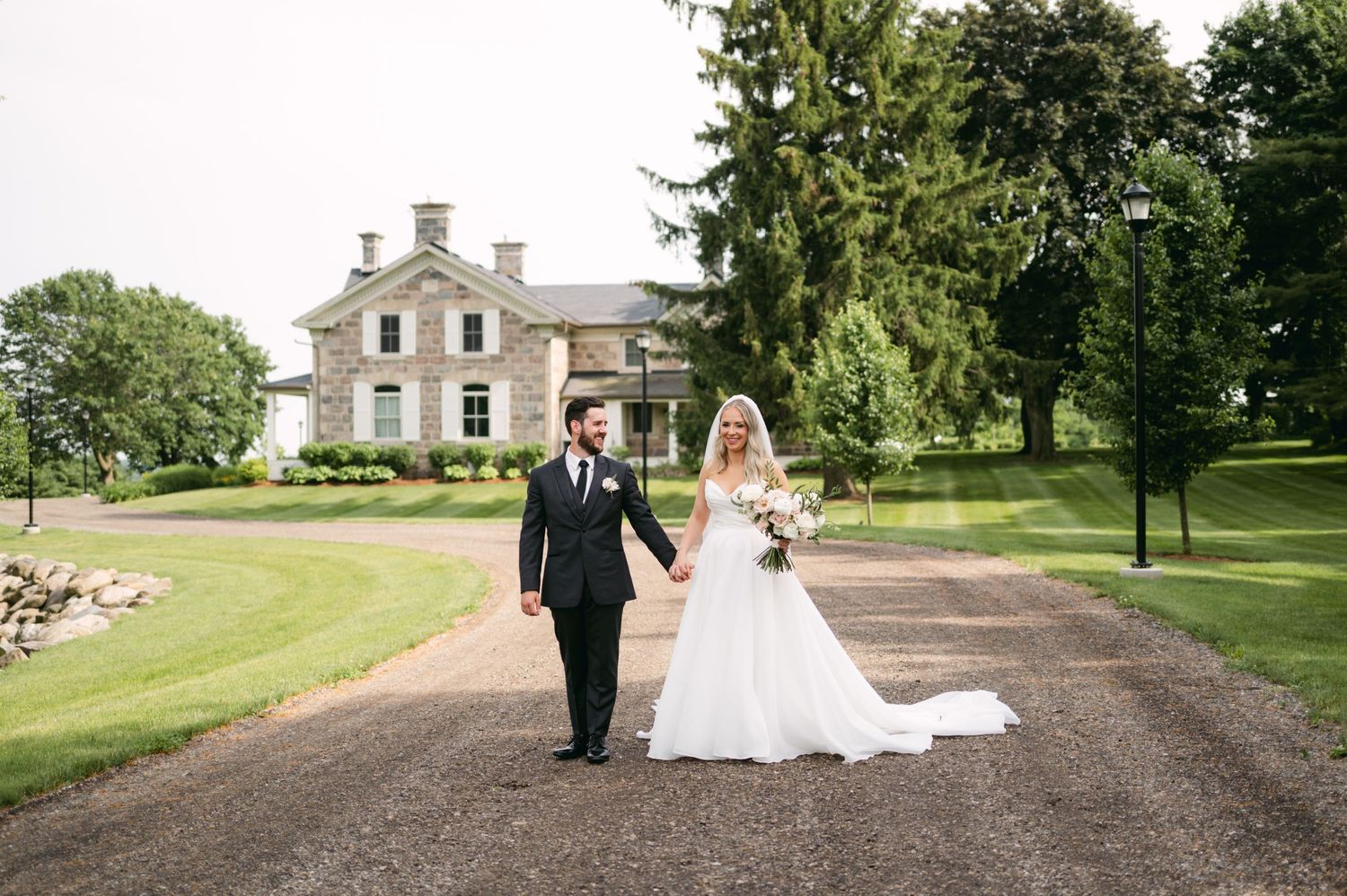 A wedding couple walks along a gravel path leading to a grand historic mansion with manicured grounds.