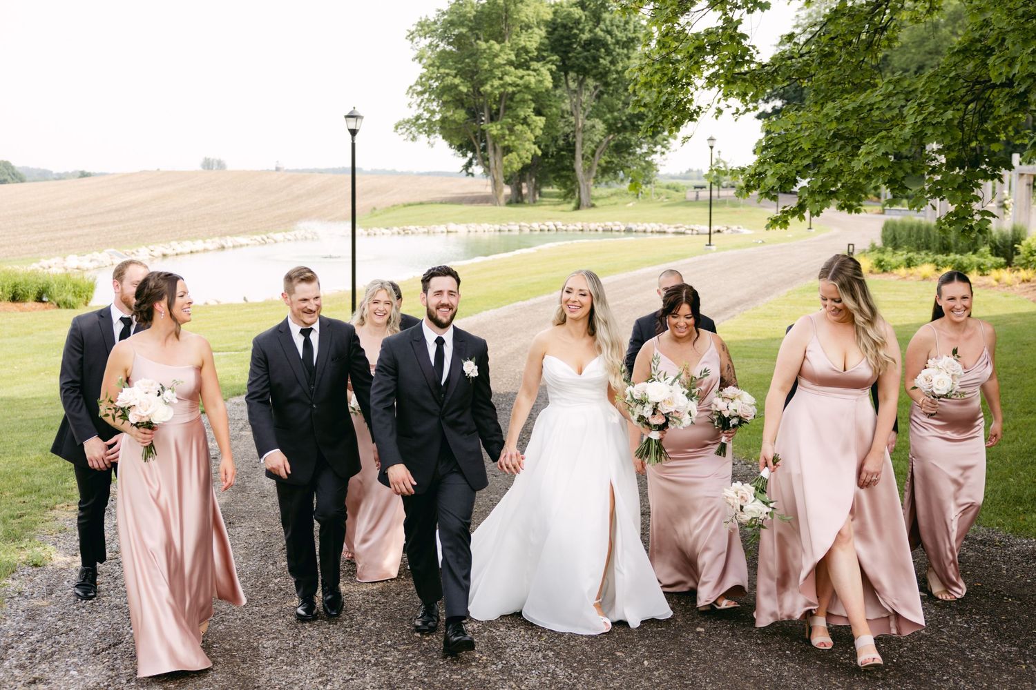 Wedding party in blush pink dresses and black tuxedos poses together outdoors.