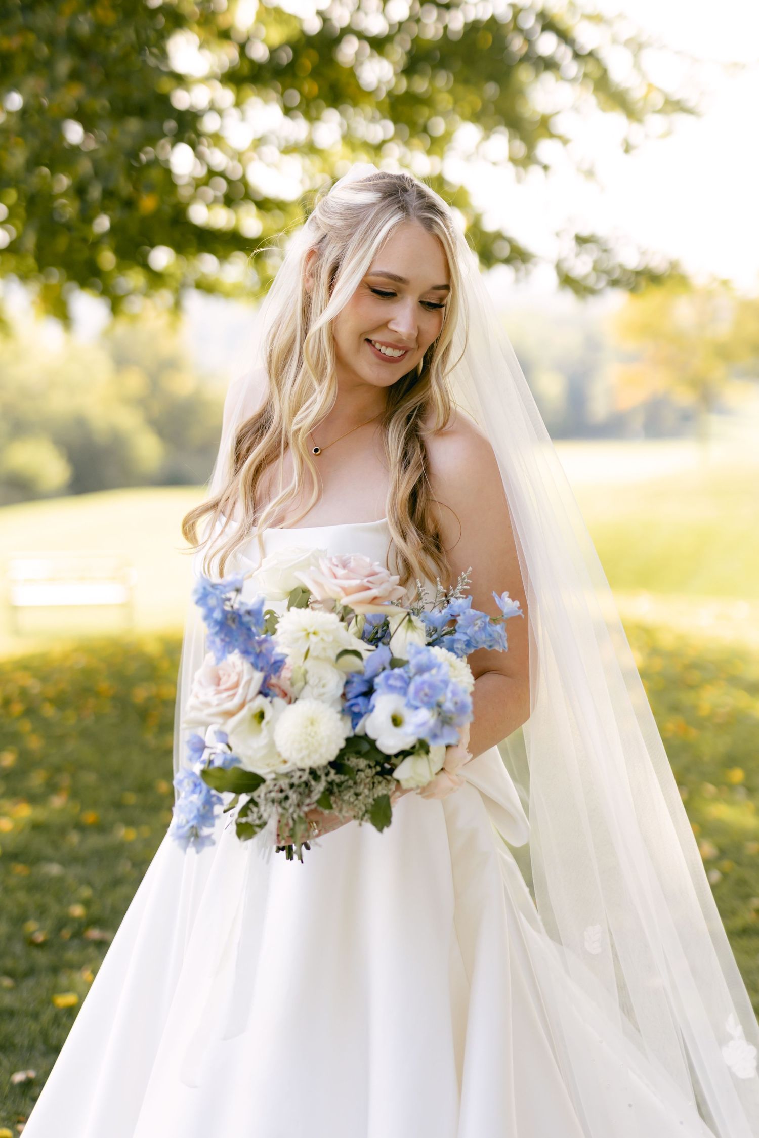 Bride in white gown holds blue and white floral bouquet while smiling outdoors.